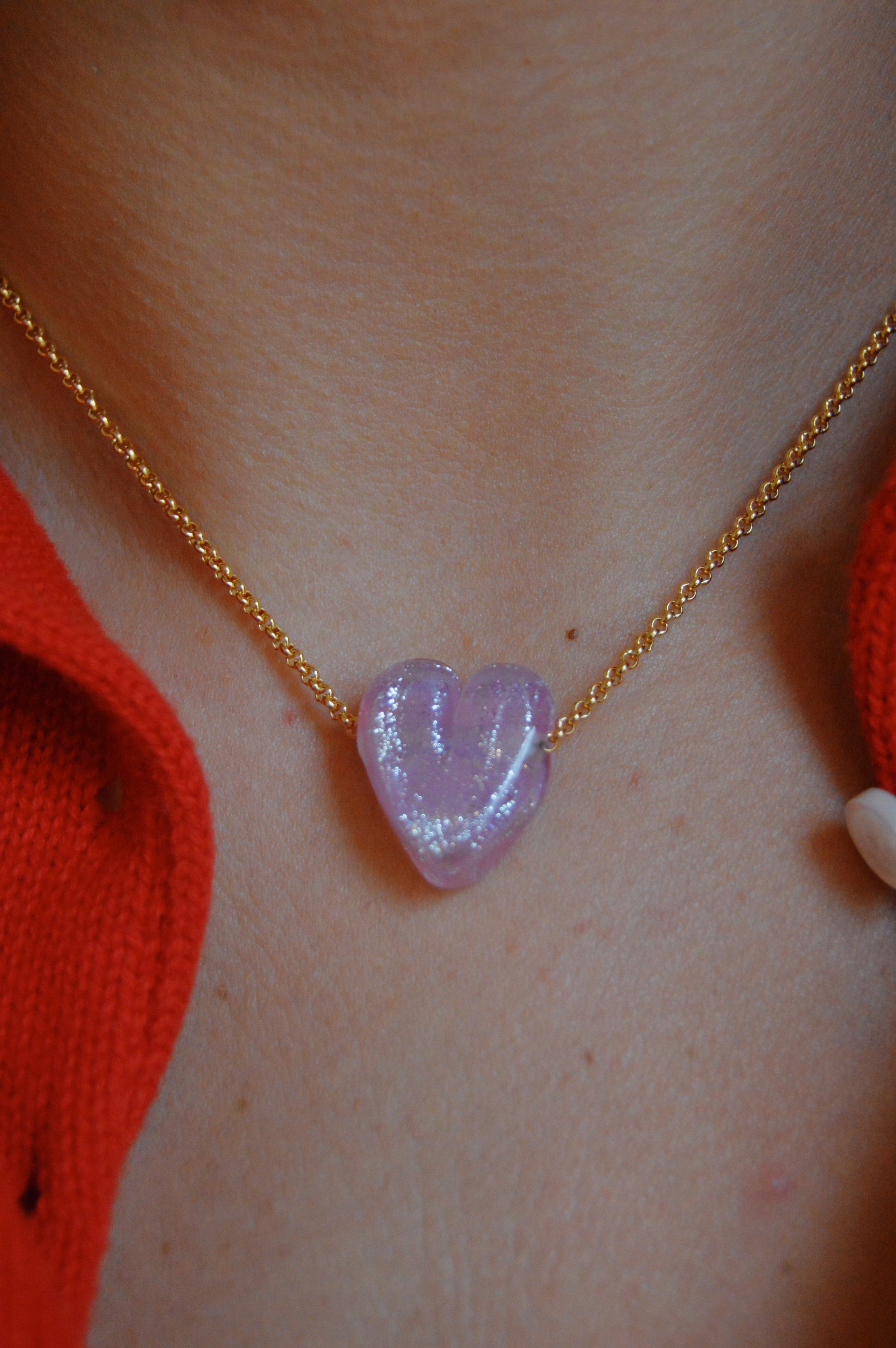 A close-up of a Glass Heart Necklaces- Pinks: a gold-filled chain with a handmade pink lampwork glass heart pendant, worn around the neck with a red garment.
