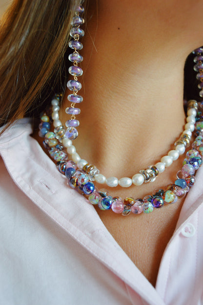 A woman in a light pink shirt showcases the Donut Drops - Shimmer Lavender: layered necklaces with pearls, colorful beads, 14k gold filled accents, and matching beaded earrings featuring handblown glass beads.