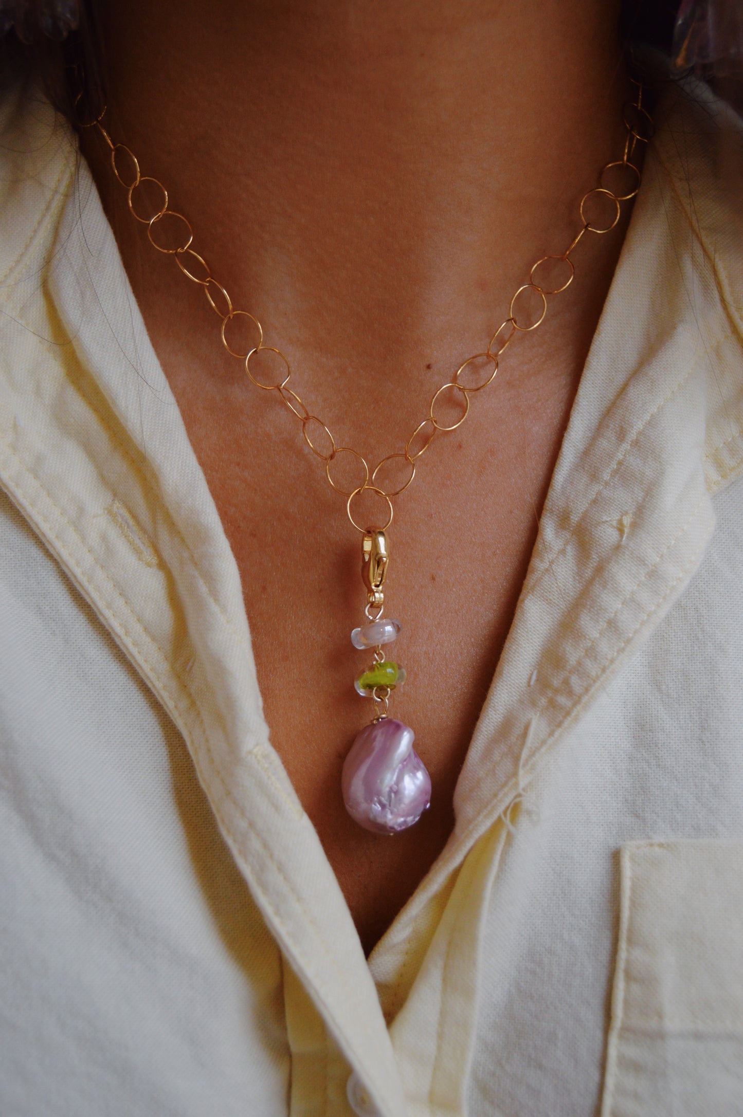 The Glass and Lavender Pearl Charm features a gold chain necklace with a cultured freshwater pearl pendant and handmade green and lavender lampwork glass beads, styled over a cream-colored button-up shirt.