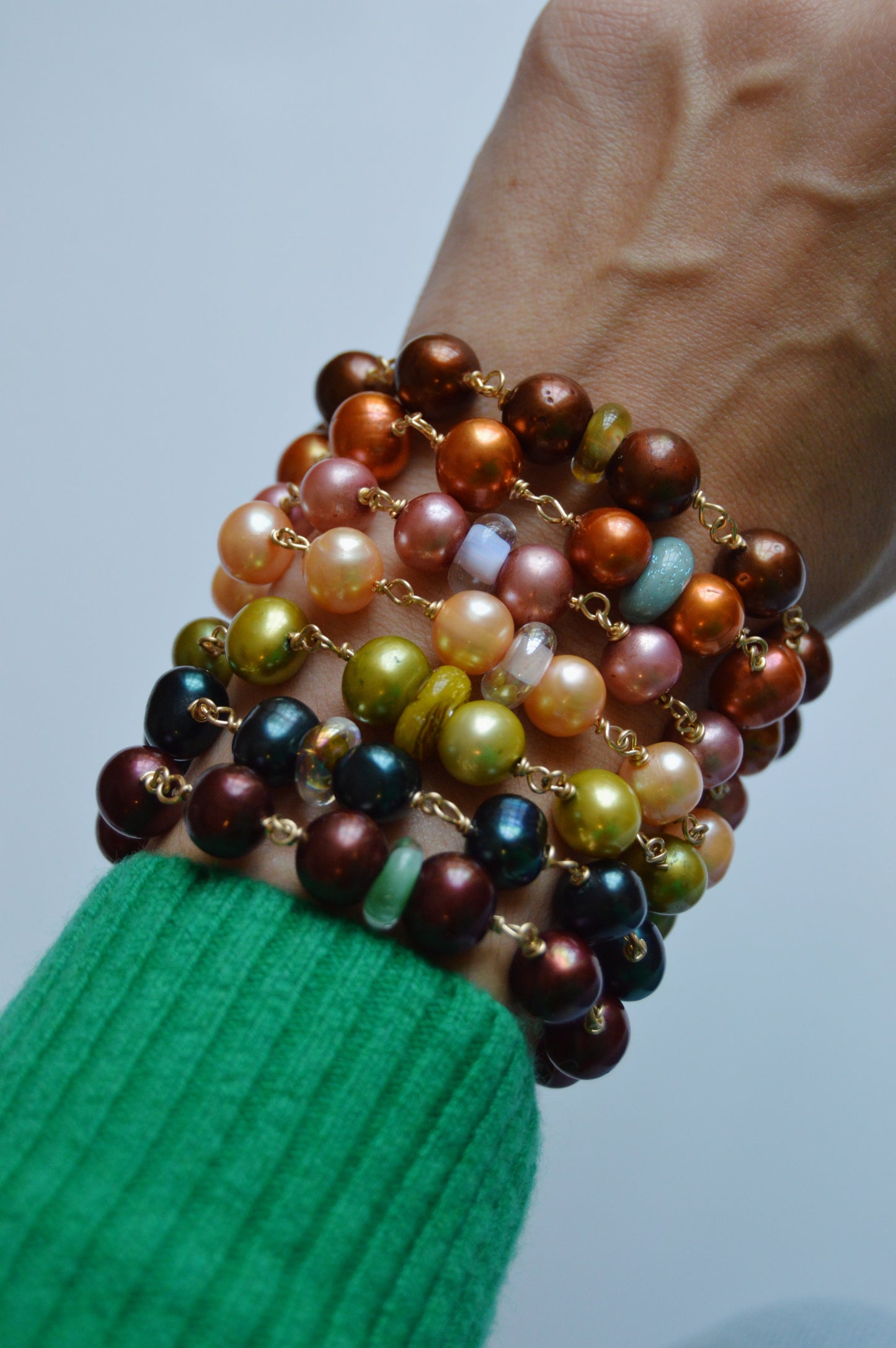 A hand in a green sleeve displays the Pearl and Glass Bracelet, featuring handmade lampwork glass beads and cultured freshwater pearls in brown, orange, gold, green, and maroon hues stacked along the wrist against a plain background.
