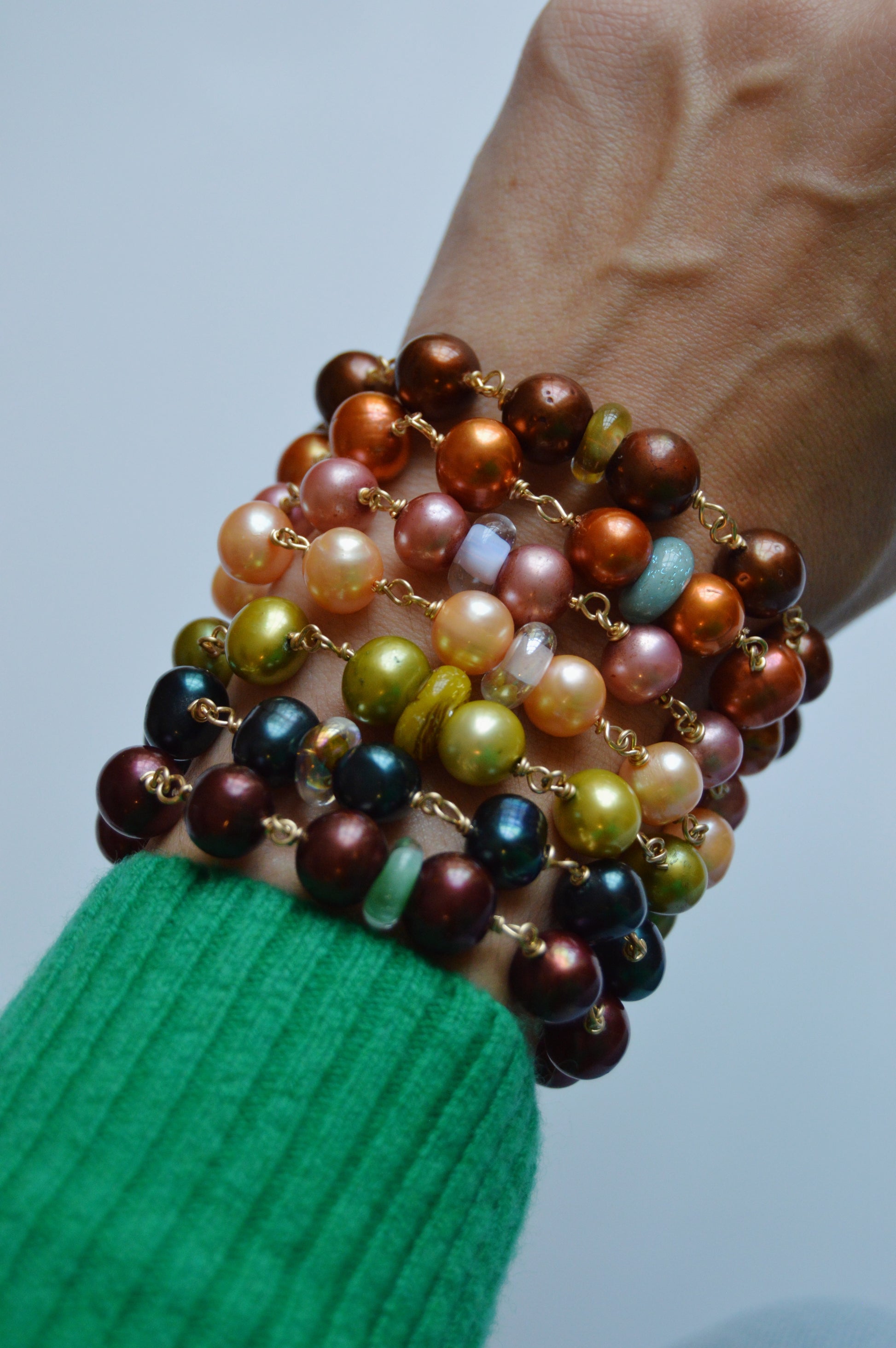 A hand in a green sleeve displays the Pearl and Glass Bracelet, featuring handmade lampwork glass beads and cultured freshwater pearls in brown, orange, gold, green, and maroon hues stacked along the wrist against a plain background.