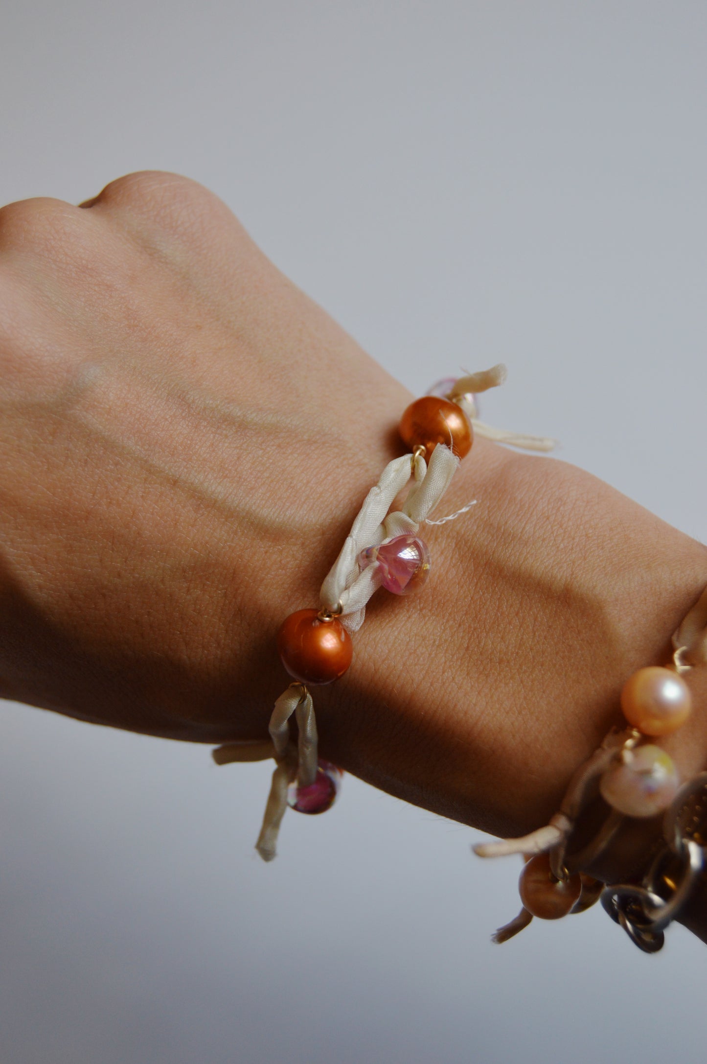 A close-up of a wrist wearing the handmade Silk and Pearl Bracelet – Shimmer Copper, featuring orange and pink beads woven with cream silk ribbon. Another freshwater pearl bracelet from the handcrafted set is also partly visible.