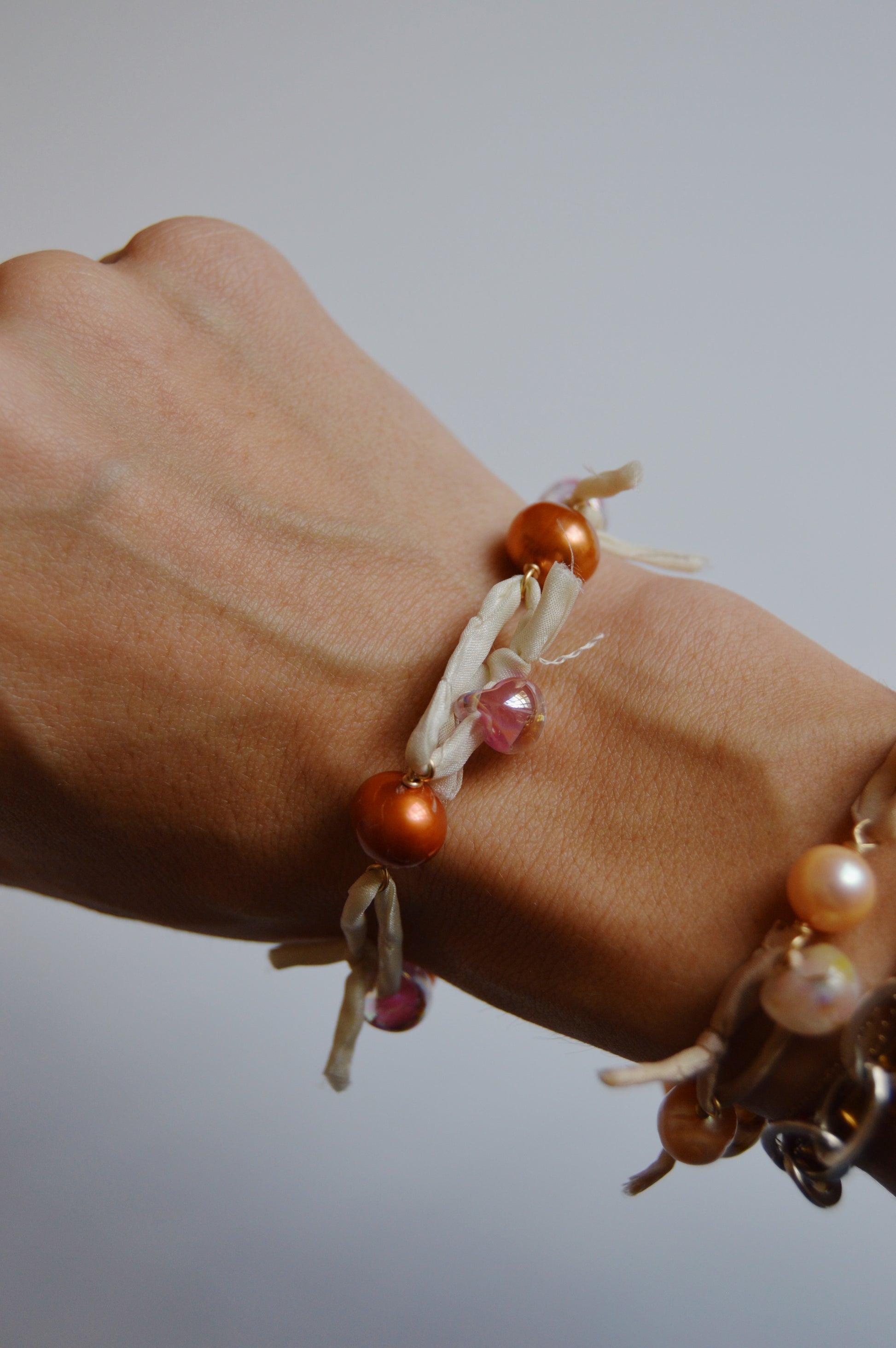 A close-up of a wrist wearing the handmade Silk and Pearl Bracelet – Shimmer Copper, featuring orange and pink beads woven with cream silk ribbon. Another freshwater pearl bracelet from the handcrafted set is also partly visible.