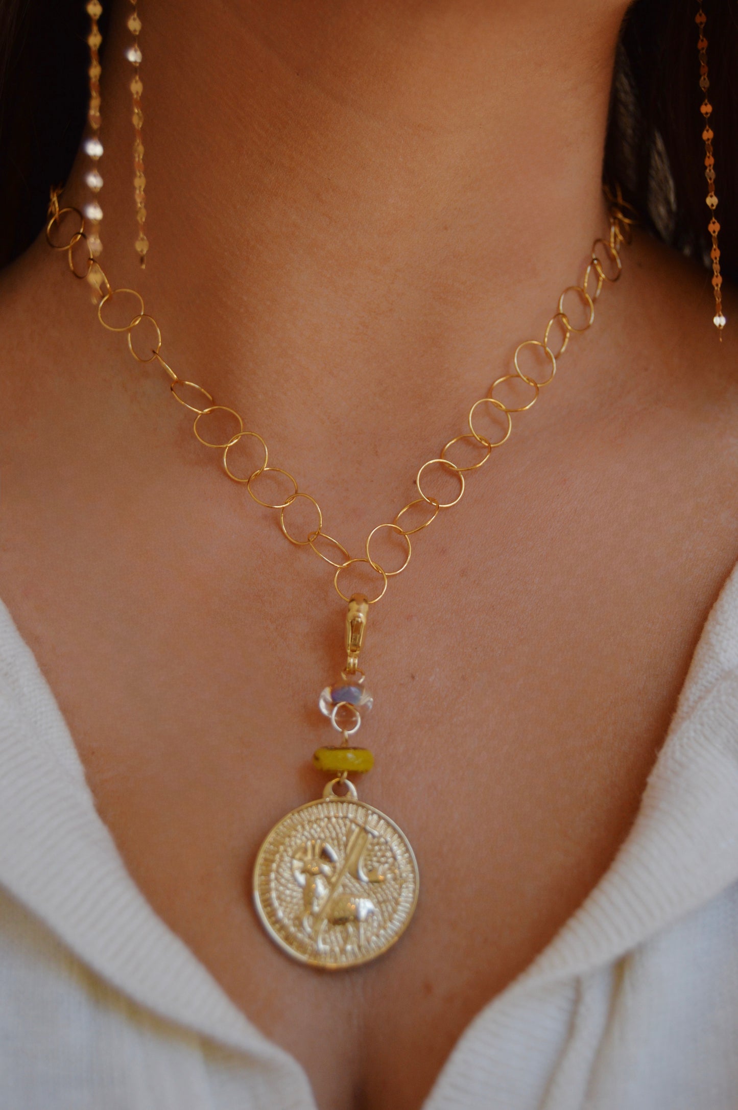 A close-up of a person wearing the Lamb of God Charm, an artisan jewelry gold chain necklace with a large ornate medallion pendant, dangle accents, and 14k gold-filled findings over a white V-neck top.