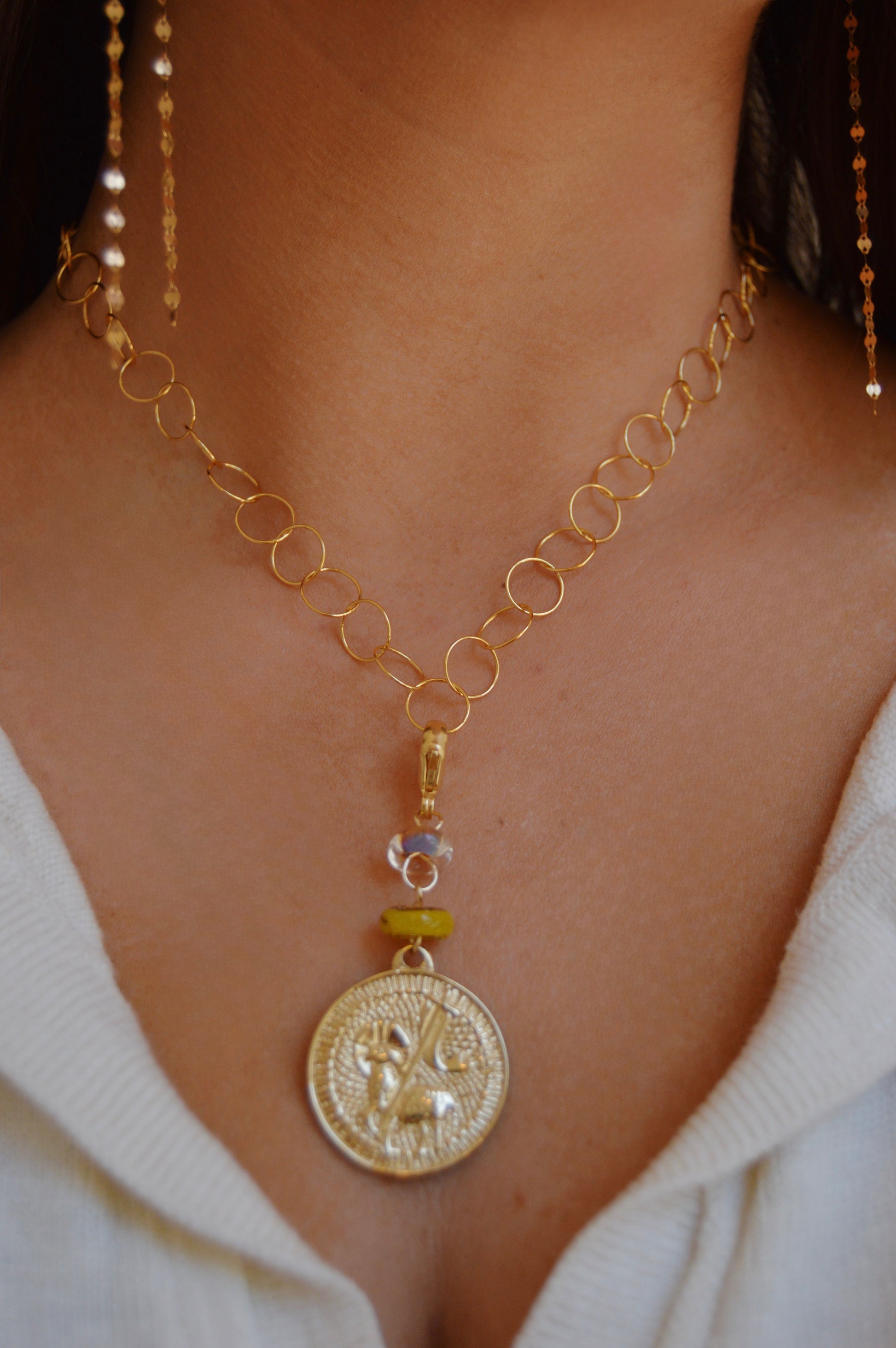 A close-up of a person wearing the Lamb of God Charm, an artisan jewelry gold chain necklace with a large ornate medallion pendant, dangle accents, and 14k gold-filled findings over a white V-neck top.