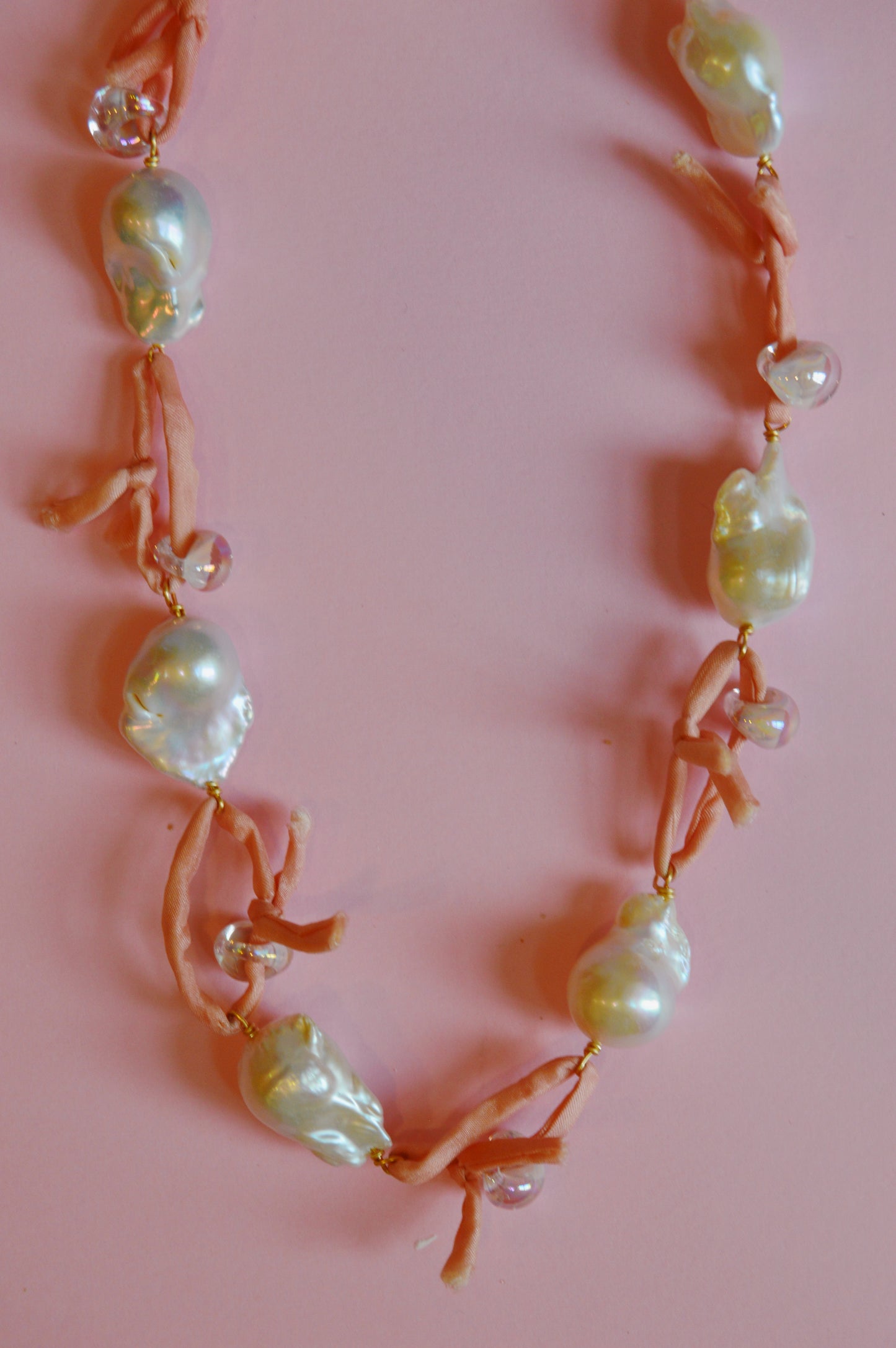 The Pink Silk and Pearl Layer necklace, featuring large cultured freshwater pearls, small clear beads, and soft pink fabric ties, is elegantly arranged in a loose curve on a light pink background.