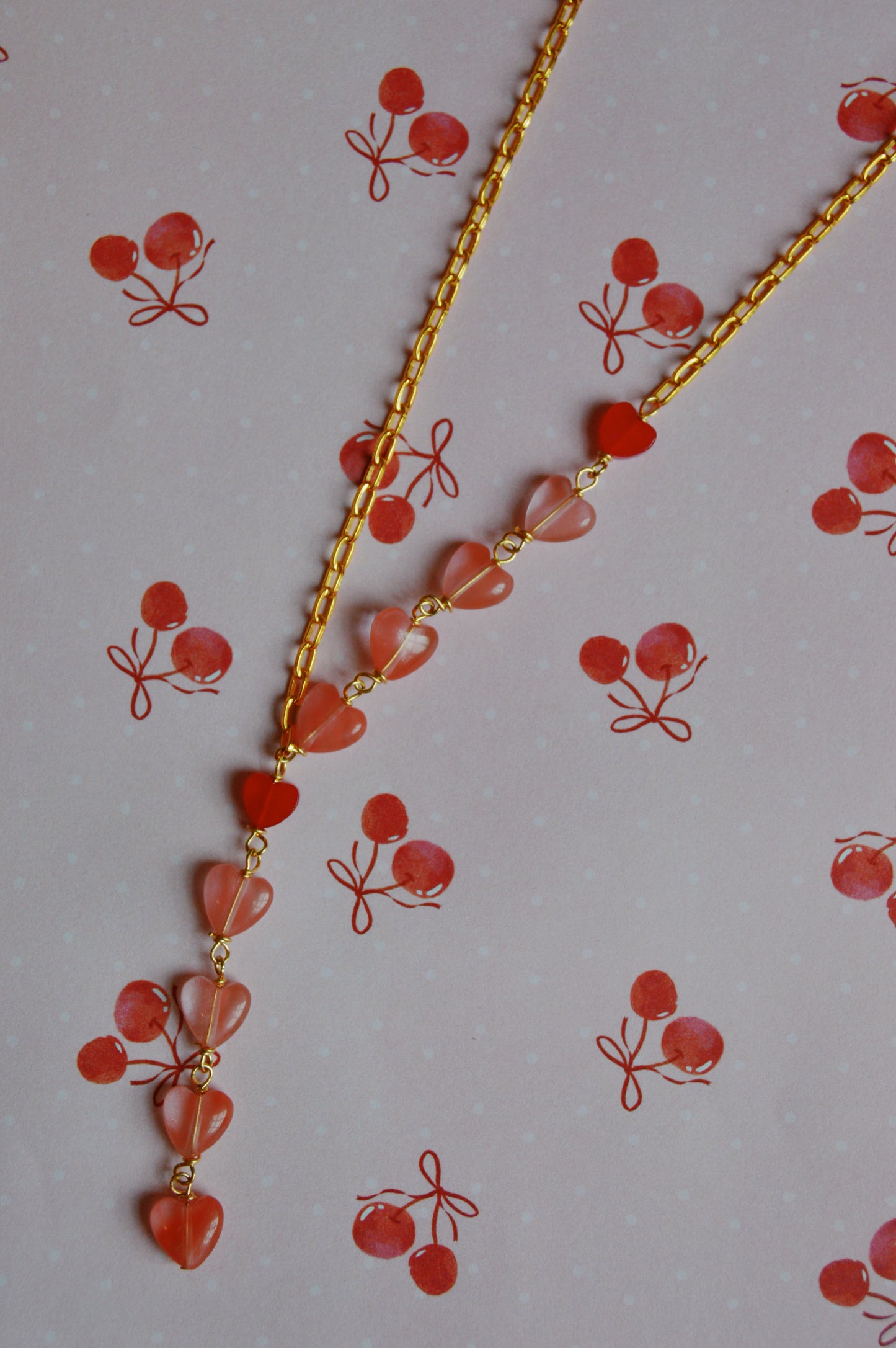 The Valentine Gemstone Heart Lariat, a gold chain necklace with small pink heart-shaped beads, is displayed on a pink background adorned with cherry illustrations.