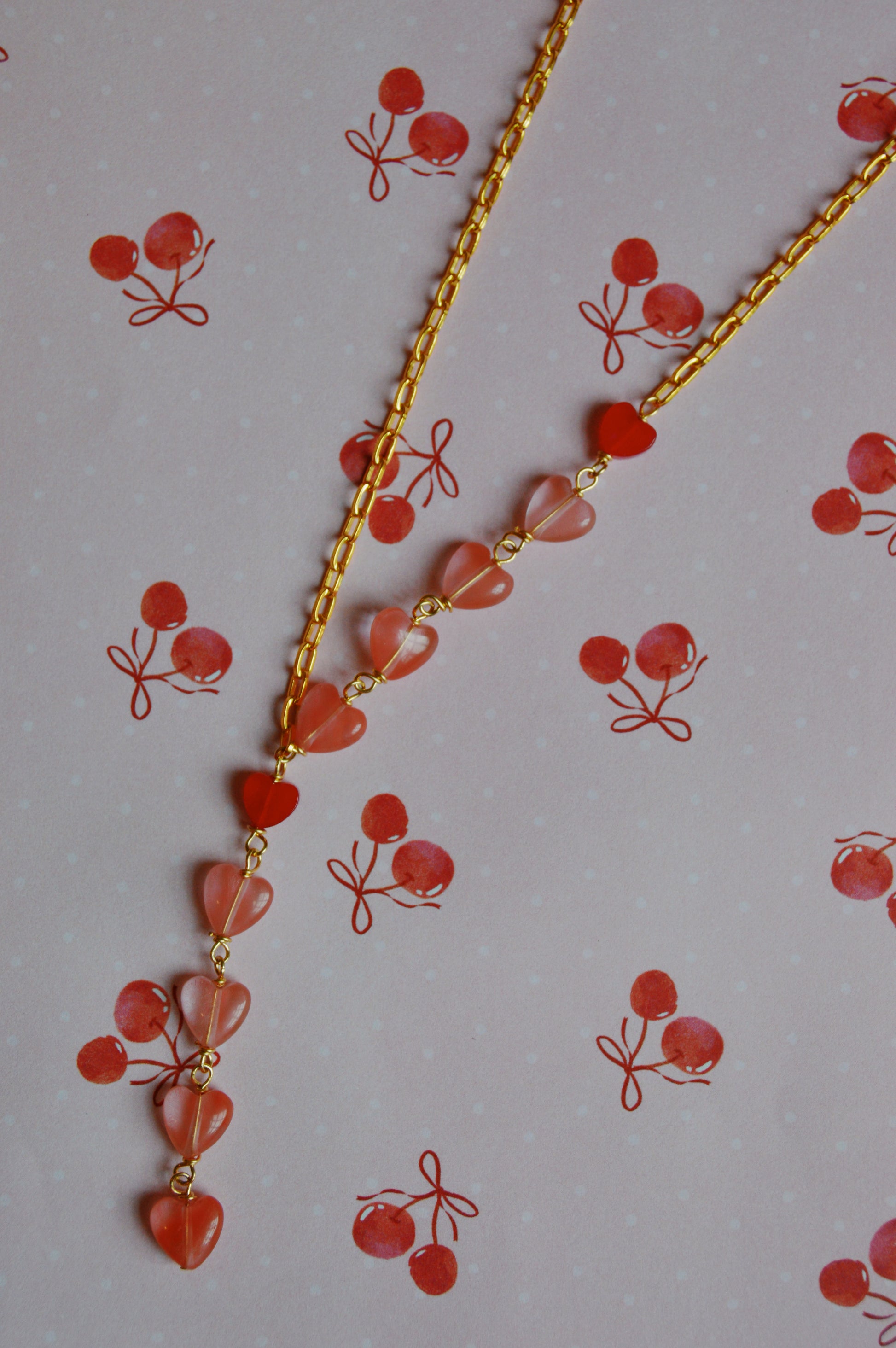 The Valentine Gemstone Heart Lariat, a gold chain necklace with small pink heart-shaped beads, is displayed on a pink background adorned with cherry illustrations.