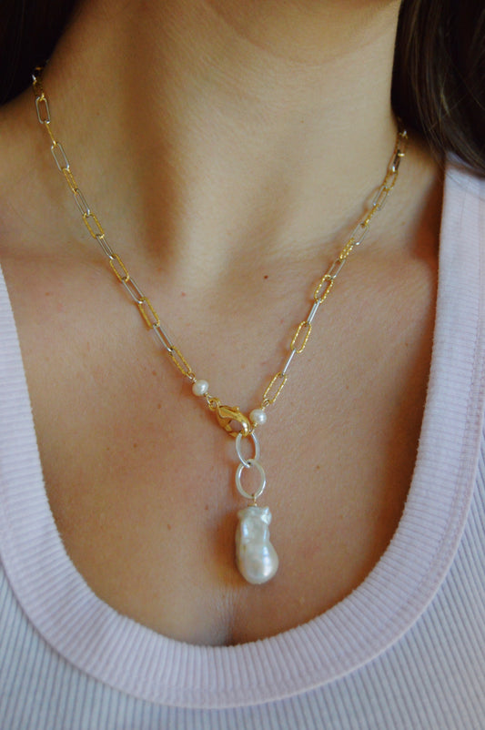 A close-up of a woman wearing the Showstopper Baroque Pearl mixed metal chain necklace with a large baroque pearl clasp pendant, paired with a light pink ribbed top.