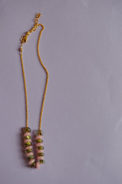 The Fall Lots of Donuts Layer, a gold chain necklace with two pendants adorned with alternating pink, green, and gray handblown glass beads, is displayed on a plain light purple background.