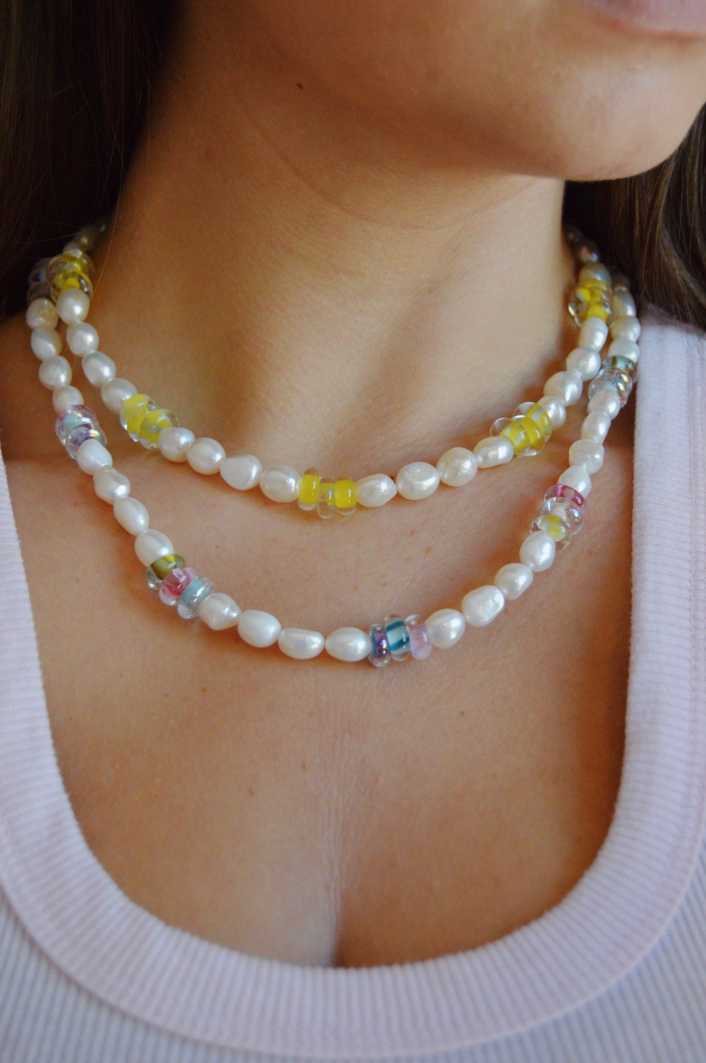 A woman in a white top wears the Glass Donut and Pearl Layer necklace, featuring cultured freshwater pearls and handblown glass beads in yellow, pink, blue, and clear. The double strand sits elegantly on her collarbone.