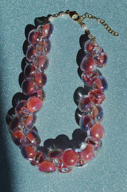 The Grand Bubble Glass Necklace-Shimmer Pink features translucent, glossy glass beads with pink centers, freshwater pearls, a gold clasp, and extension chain, displayed on a sparkling teal surface with a bold shadow.