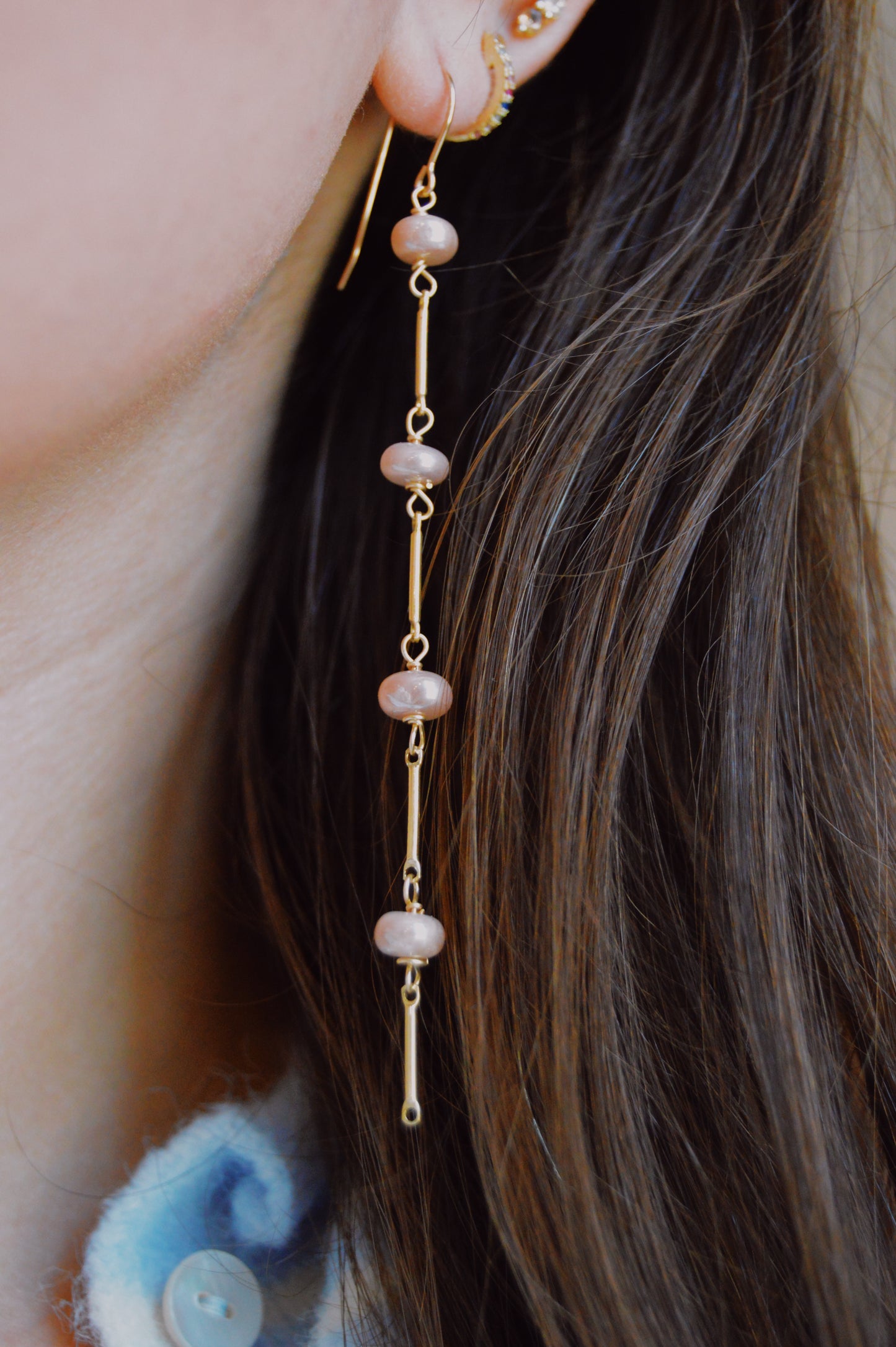 A close-up of a person wearing the Moonstone Dot Drops—a long gold-filled earring with four pink beads on delicate rods—beside brown hair and paired with a blue and white collared shirt.