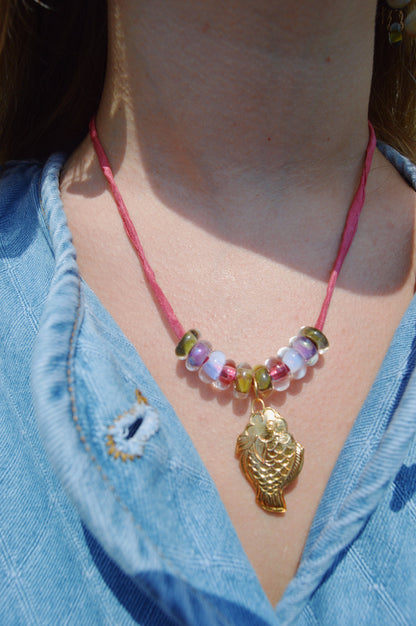 Close-up of a person wearing the Cloisonne Fish Layer - Berry necklace, featuring handblown glass beads and a gold seashell pendant on a pink cord, paired with a blue denim shirt with a frayed buttonhole.