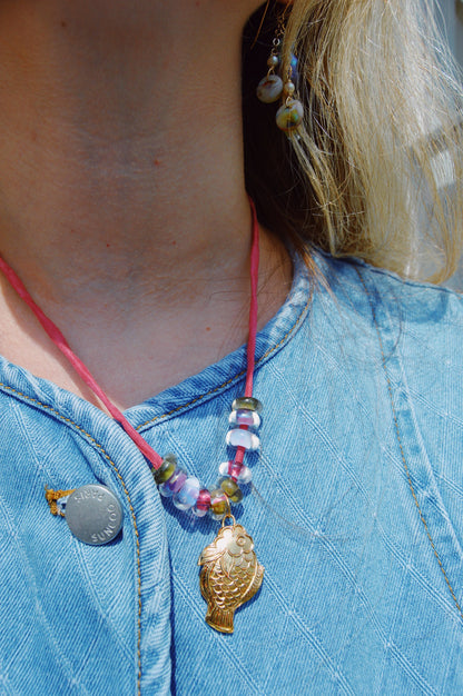 A person wears a blue quilted top with the Cloisonne Fish Layer - Berry necklace featuring colorful beads and a cloisonné fish pendant, a round silver pin, dangling earrings with handblown glass beads and shells. Blonde hair shows partially.