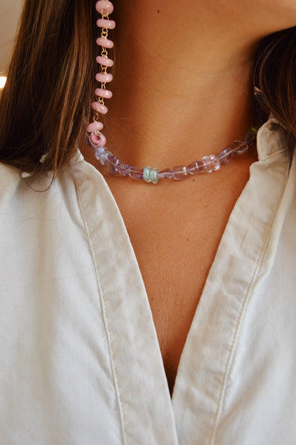 A close-up shows a person with brown hair wearing a white shirt, the Amethyst Dreams Layer purple handblown glass bead necklace, and a pink beaded earring. The focus is on their neck and chest.