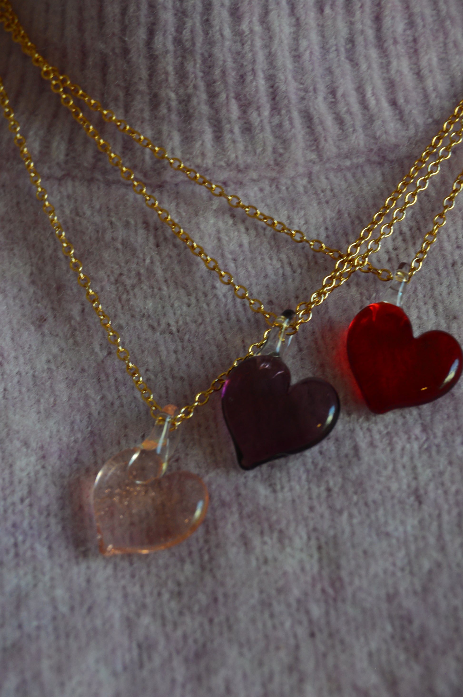 Three Flat Glass Heart Layer necklaces—gold-filled chains with 14k gold pink, purple, and red handblown glass heart pendants—are displayed on a light purple knit sweater.