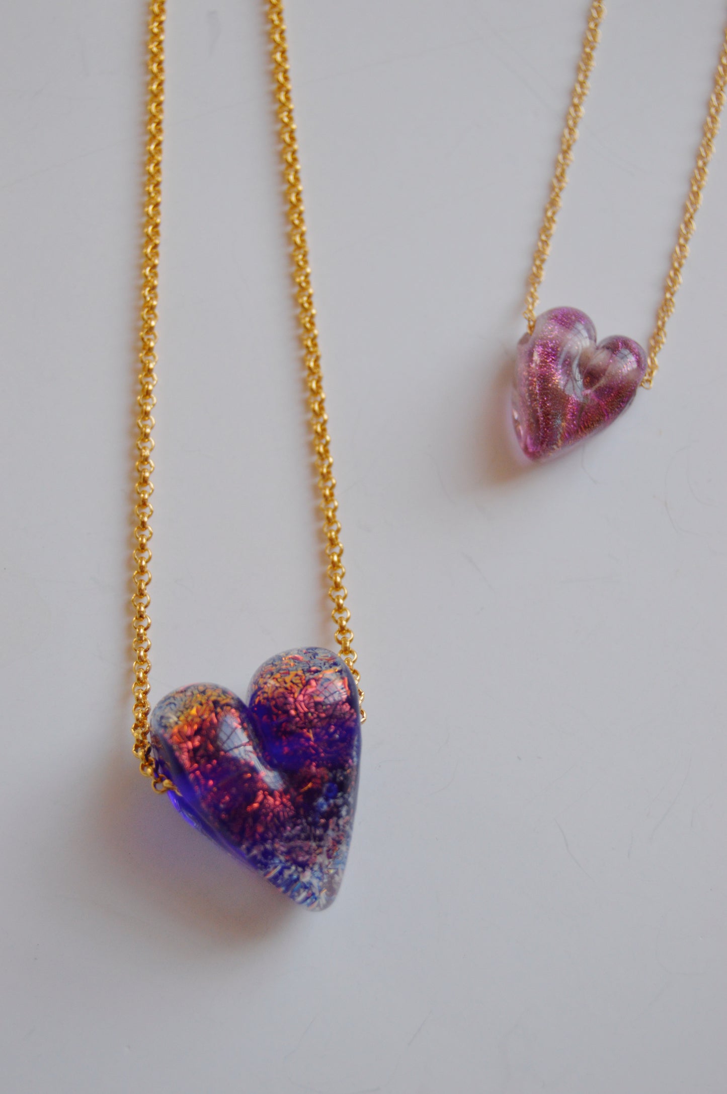 The Glass Heart Necklaces- Purples set features two gold-filled chain necklaces: one with a handmade speckled blue lampwork glass heart, the other with a shimmery light purple heart, both elegantly displayed on a white background.