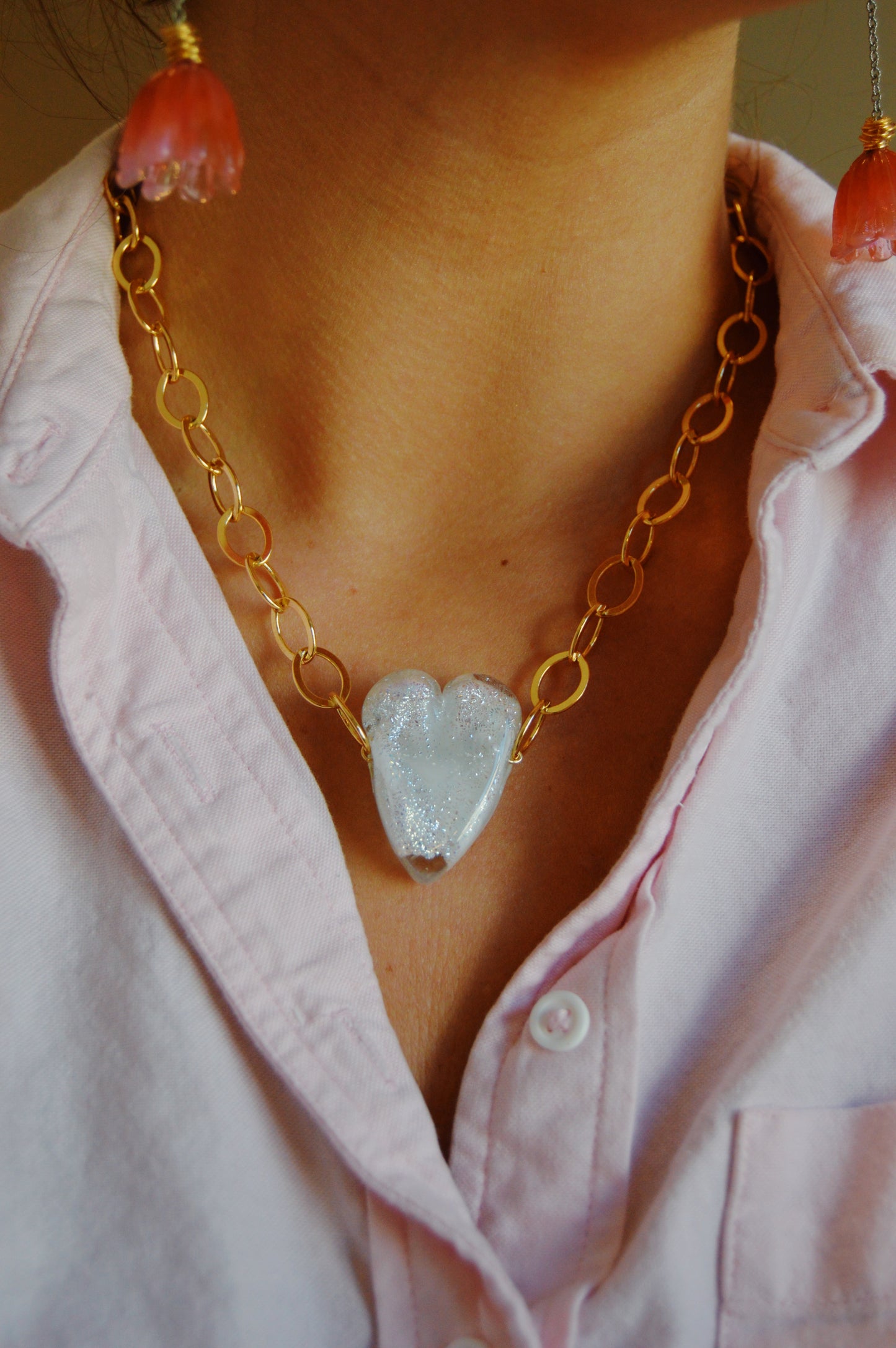 A person wears the "White Glass Heart on Chunky Gold Filled Chain," featuring a large handmade glass heart pendant, paired with a light pink button-up shirt and partially visible coral tassel earrings.