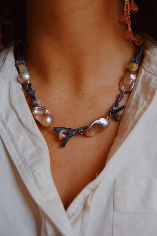 A woman wears a white collared shirt and the Silk and Pearl Layer-Blueberry Puka necklace, featuring hand-knotted irregular pearls on dark string. A pink stone earring is partially visible.