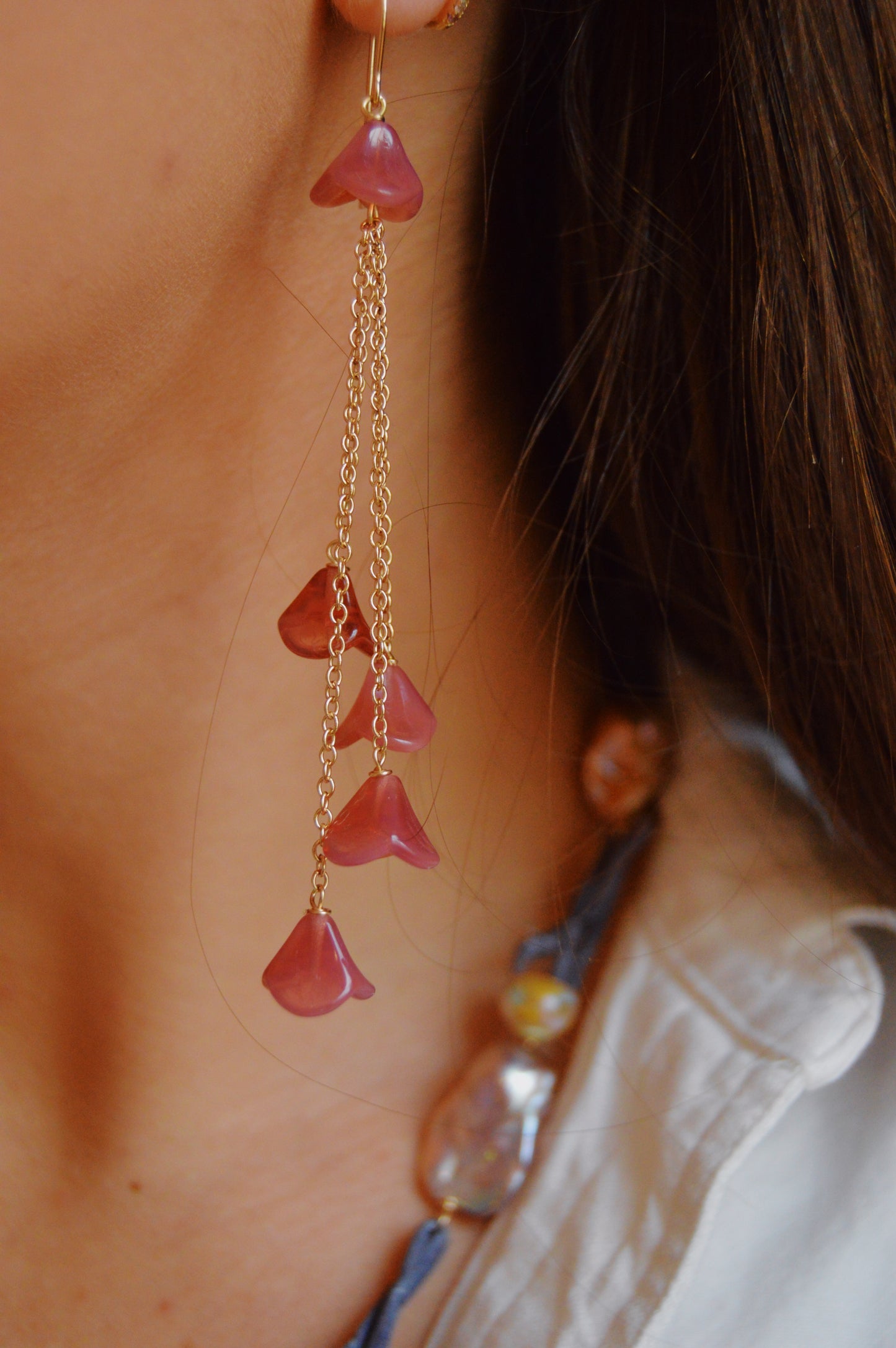 Close-up of a person wearing Bouquet Drops- Raspberry: handcrafted long, dangling earrings featuring pinkish-purple Czech Glass Tulips on 14k gold-filled chains, styled with a necklace and a white shirt.