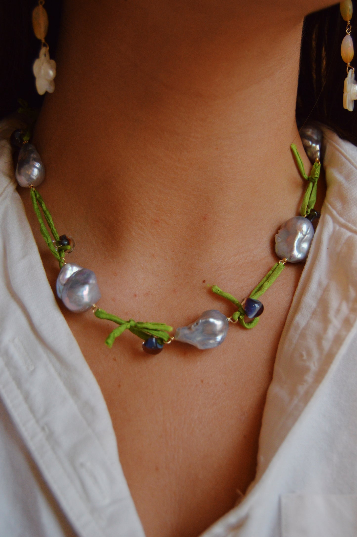 Wearing a white shirt, the person showcases the Silk and Pearl Layer- Tahitian Palm necklace, handmade with large, irregular freshwater pearls and green thread. A matching pearl earring is partly visible on the left.