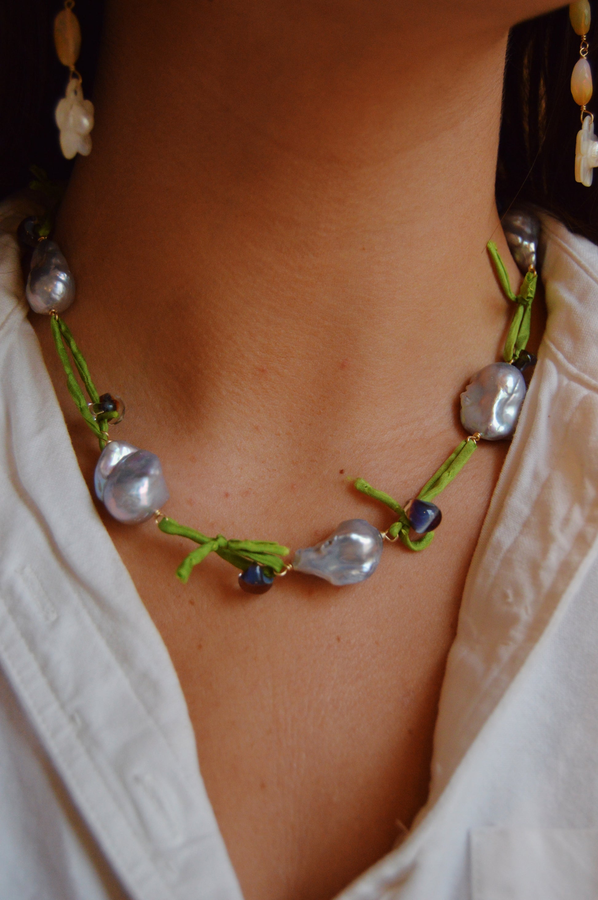 Wearing a white shirt, the person showcases the Silk and Pearl Layer- Tahitian Palm necklace, handmade with large, irregular freshwater pearls and green thread. A matching pearl earring is partly visible on the left.