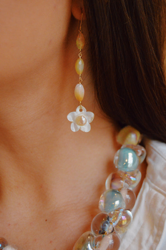 Close-up of a person wearing Opal & Pearl Flower Drops—dangling earrings with iridescent beads and a Mother of Pearl flower—paired with a chunky translucent bead necklace. The person is dressed in a white top.