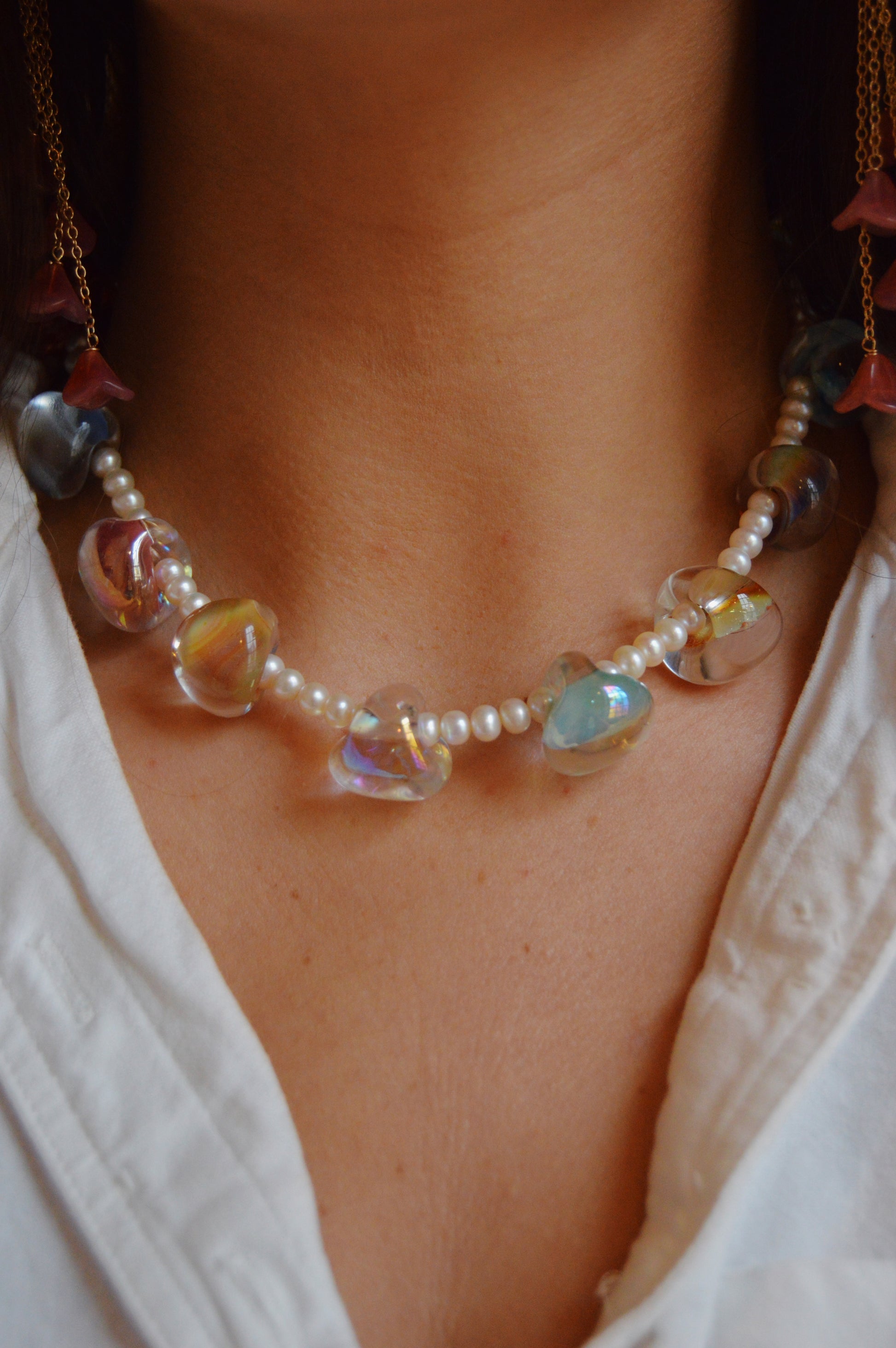 A close-up of a person wearing the Grand Bubble and Pearl Layer necklace—freshwater pearls with iridescent lampwork glass beads in pink and blue hues—styled over a white shirt, sitting gracefully at the base of the neck.