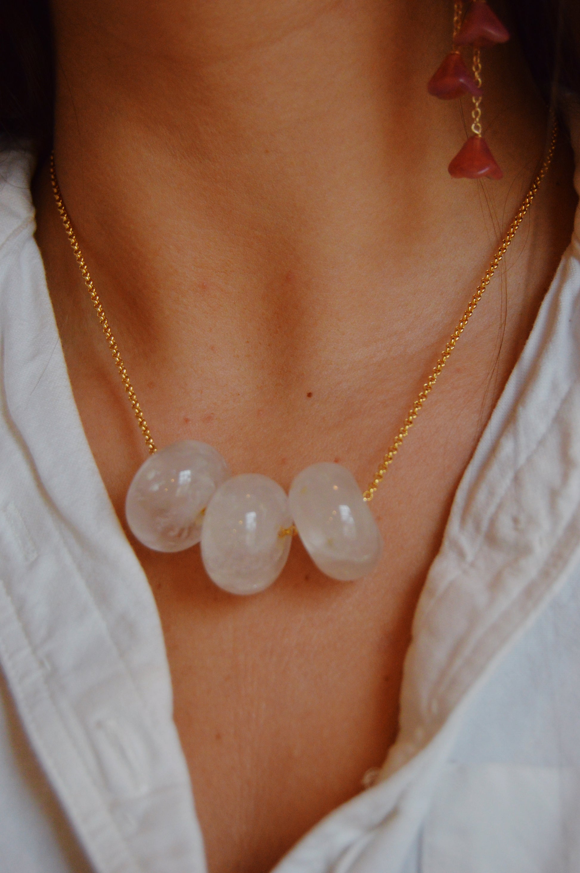 A person wears a white shirt and the Grand Donut Layer-Clear 14k gold-filled chain necklace featuring three large translucent white beads; a handcrafted earring with red beads is also partially visible.