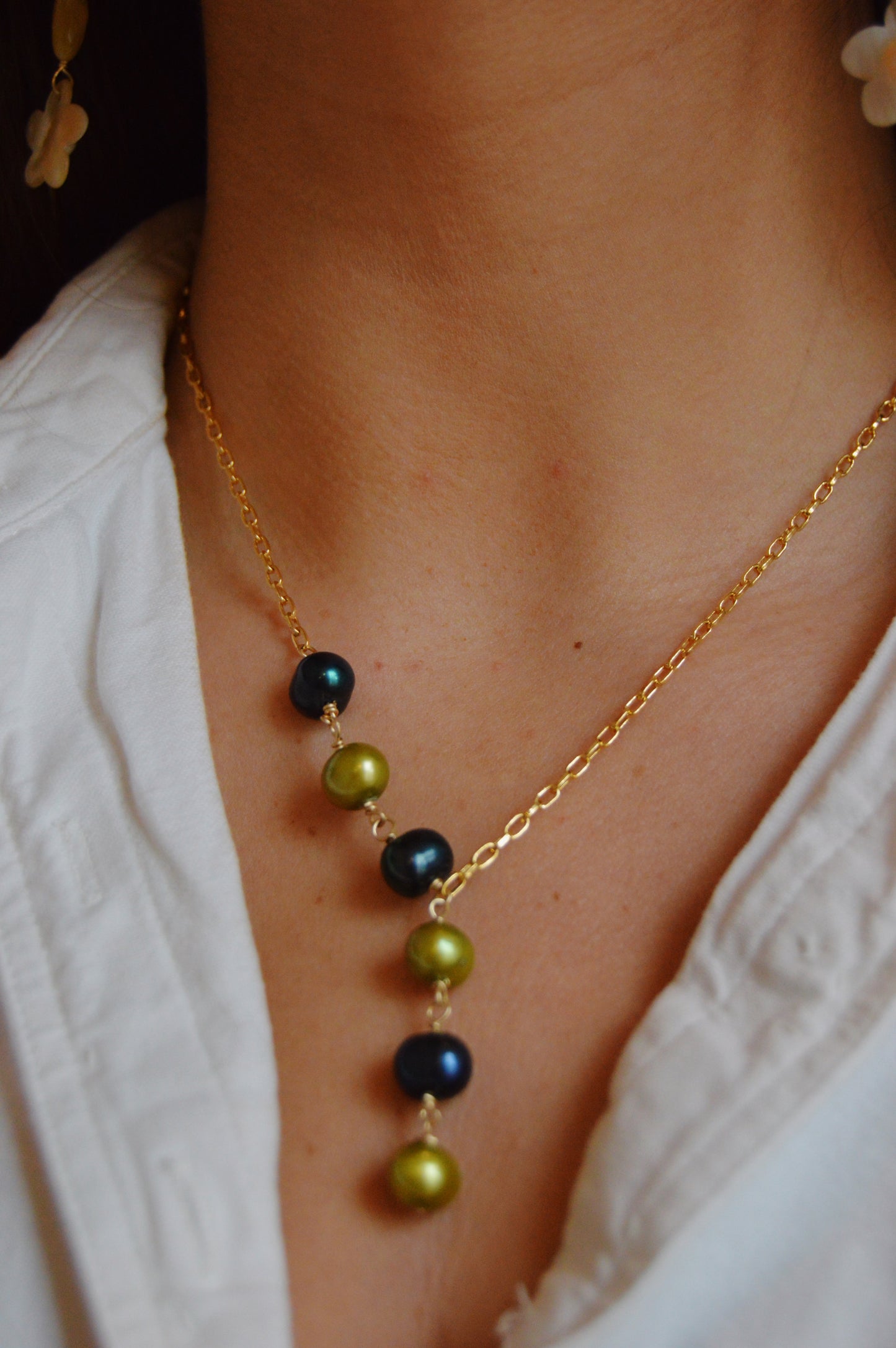 A close-up of a person wearing the Six Pearl Lariat- Blue and Green Mix, a handcrafted 14k gold-filled chain necklace with green and dark blue beads, styled with a white shirt and partially visible floral earrings.