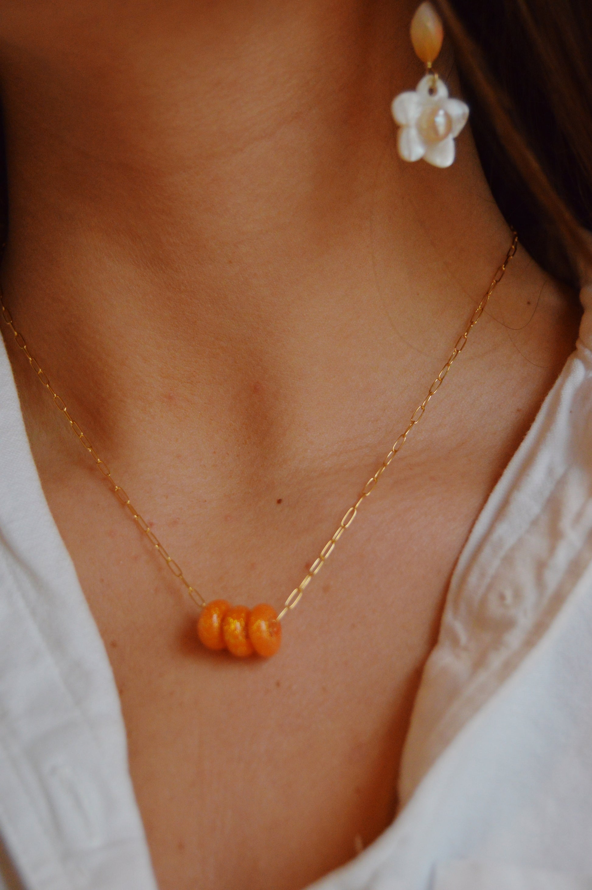 A close-up of a person wearing the Dainty Donut Layer- Sunshine: a gold-filled chain necklace with handmade lampwork glass beads, paired with a white collared shirt and a delicate pearl flower earring.