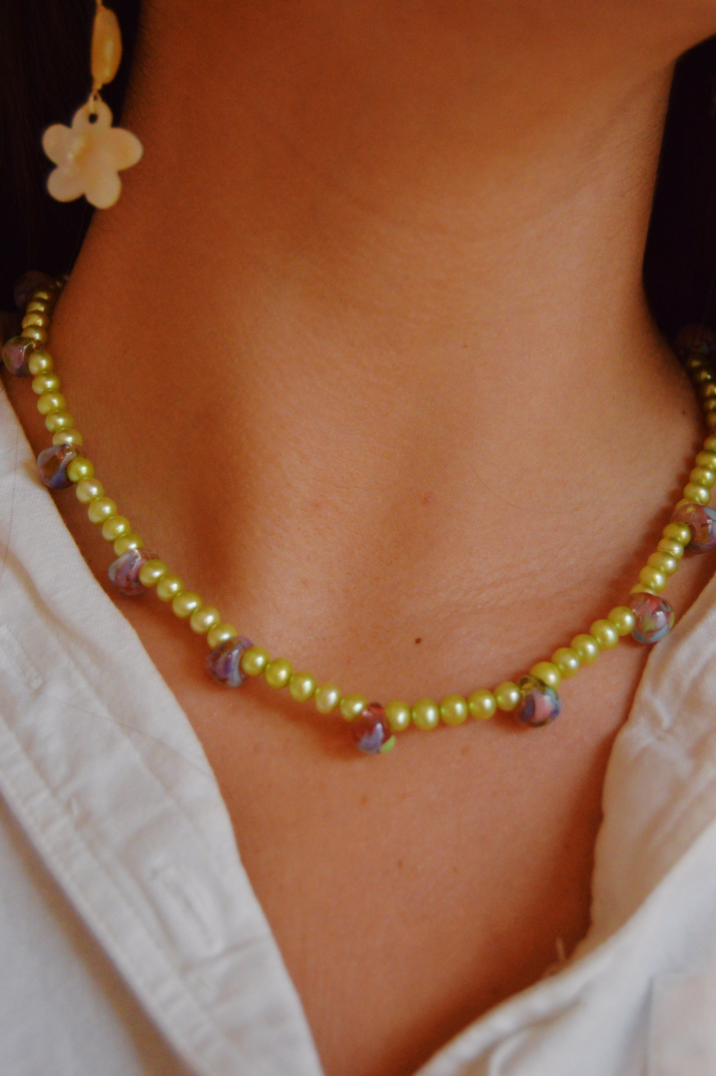 Close-up of a person wearing the Bubble and Pearl Layer, a light green beaded necklace with handmade lampwork glass beads, paired with a white flower-shaped earring and a white collared shirt.