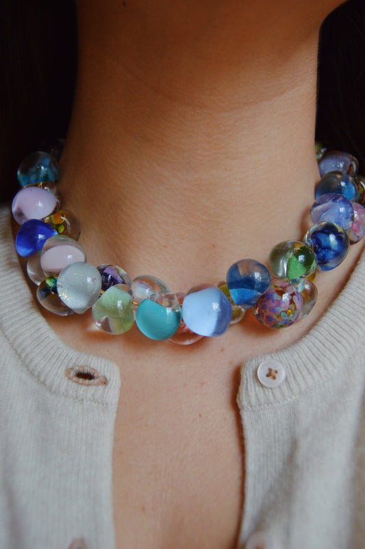 A close-up of a person wearing the Grand Bubble Necklace- Seaglass Mix, featuring large, colorful handmade lampwork glass beads. The handcrafted necklace pairs perfectly with a cream-colored buttoned cardigan, open at the collar.