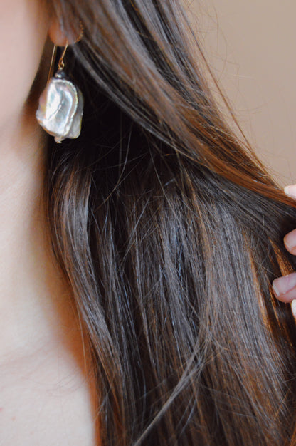 Close-up of a person with long brown hair holding a lock of hair, wearing Pearl Drops—a large, irregularly shaped Cultured Freshwater Pearl earring. This elegant piece of handcrafted jewelry stands out against a softly blurred background.