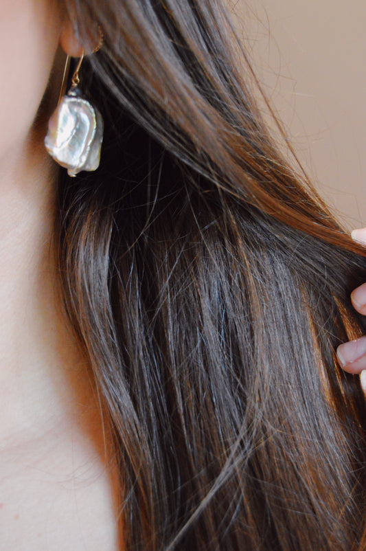 Close-up of a person with long brown hair holding a lock of hair, wearing Pearl Drops—a large, irregularly shaped Cultured Freshwater Pearl earring. This elegant piece of handcrafted jewelry stands out against a softly blurred background.