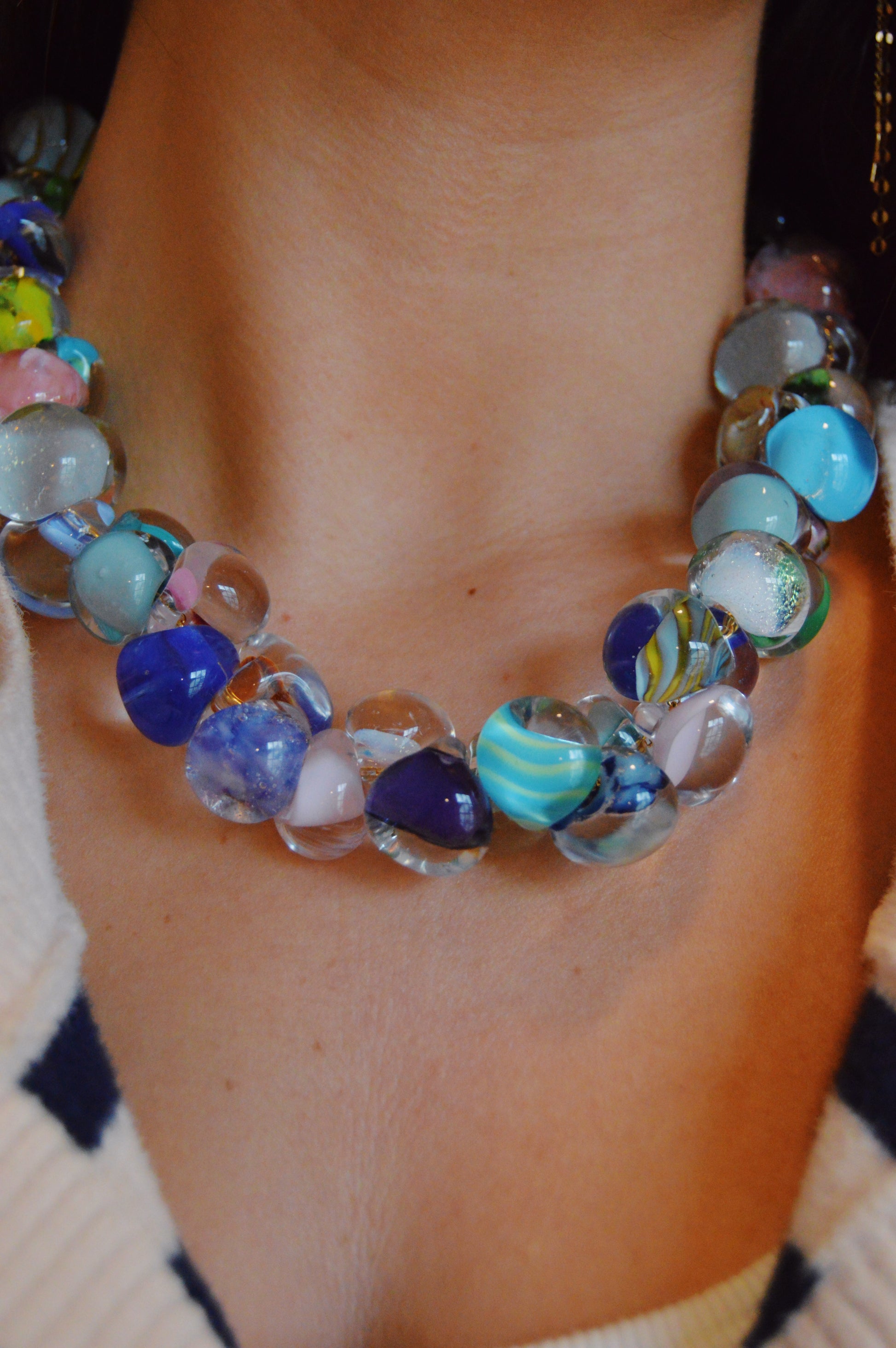 A close-up of a person’s neck showcases the Grand Bubble Glass Necklace- Poolside Mix, featuring chunky handmade lampwork beads in blue, pink, and clear tones, paired with a cream and navy shirt.