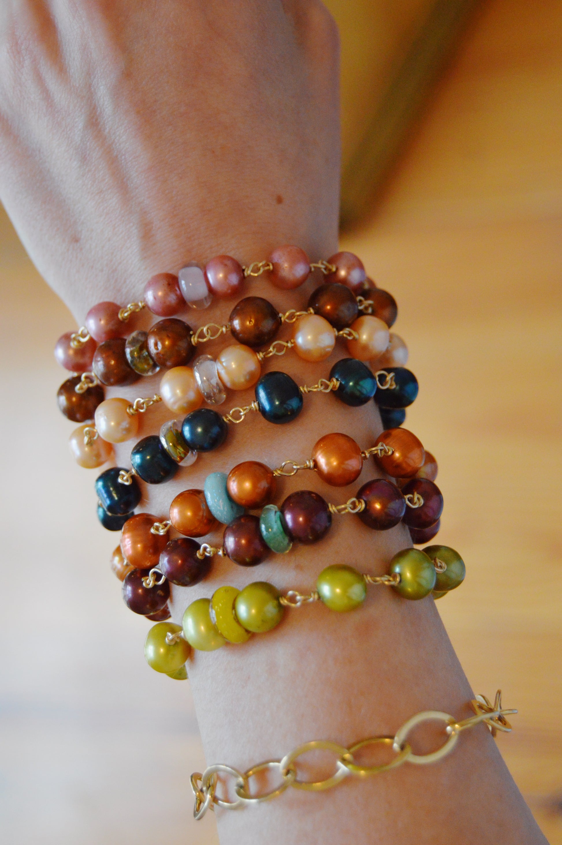 A hand wears the Pearl and Glass Bracelet, featuring colorful handmade lampwork glass beads with shiny brown, blue, yellow, green, and pink accents on a 14k gold-filled chain, set against a blurred warm-toned background.