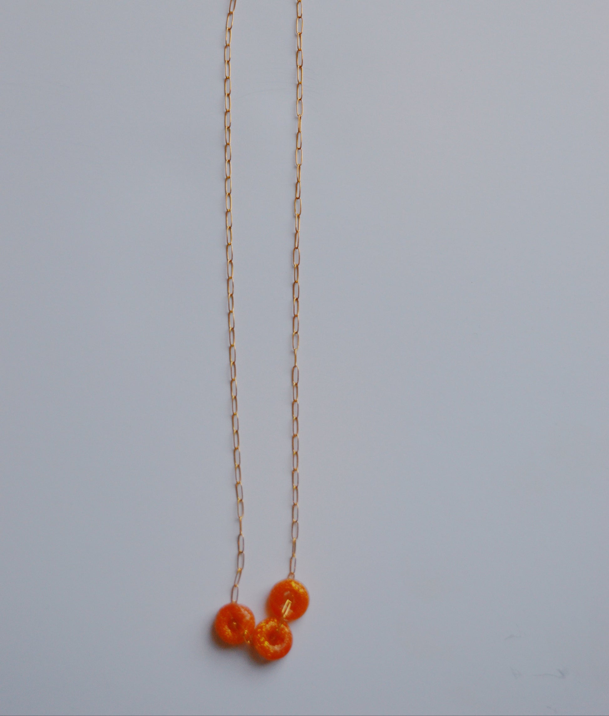 The Dainty Donut Layer- Sunshine features a delicate gold-filled chain with three small orange fuzzy pom-poms at the center, highlighting handcrafted charm against a light gray background.