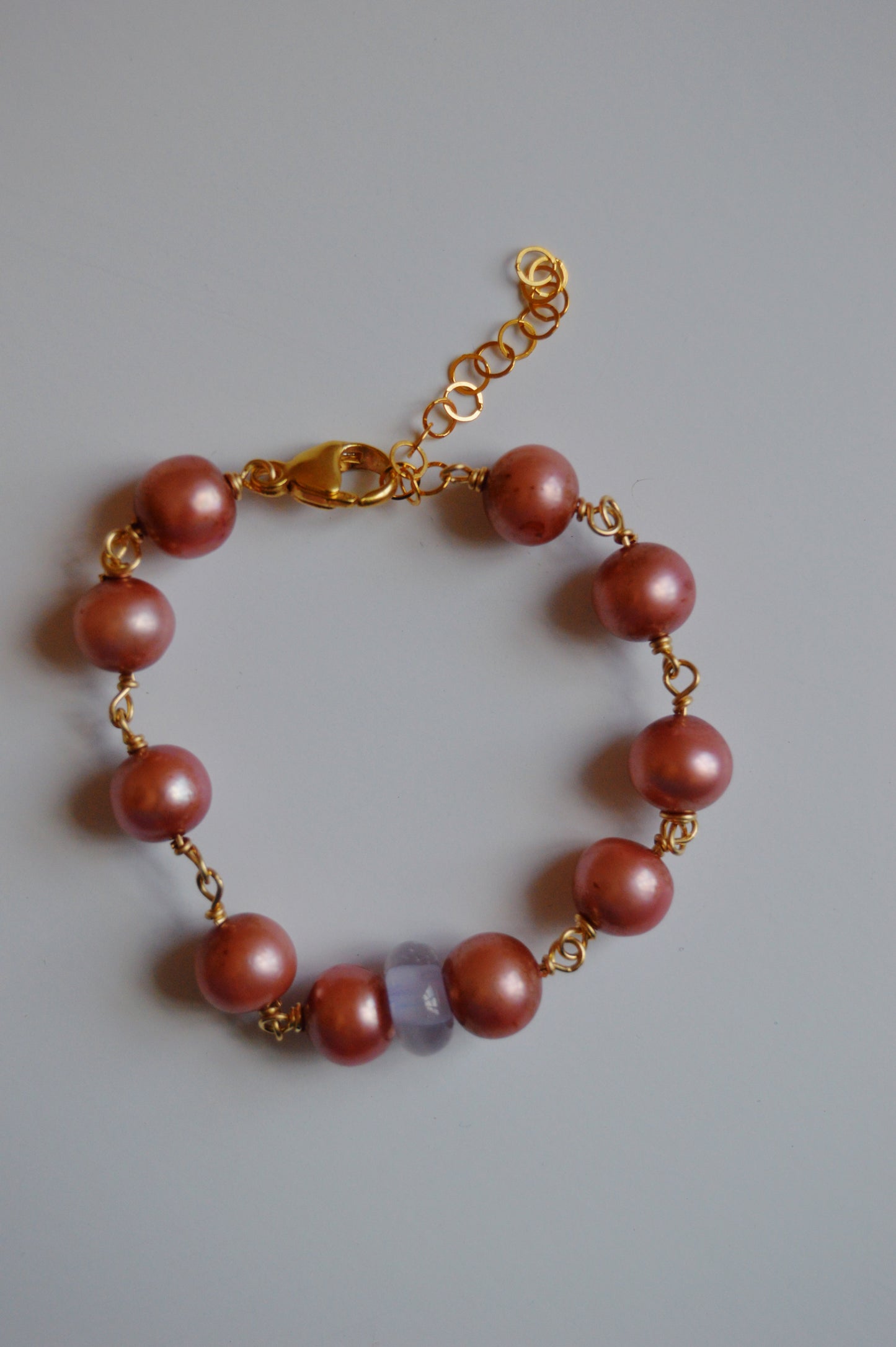 The Pearl and Glass Bracelet features pink handmade lampwork glass beads, a translucent lavender bead, and a 14k gold-filled chain with clasp, displayed on a light gray background.