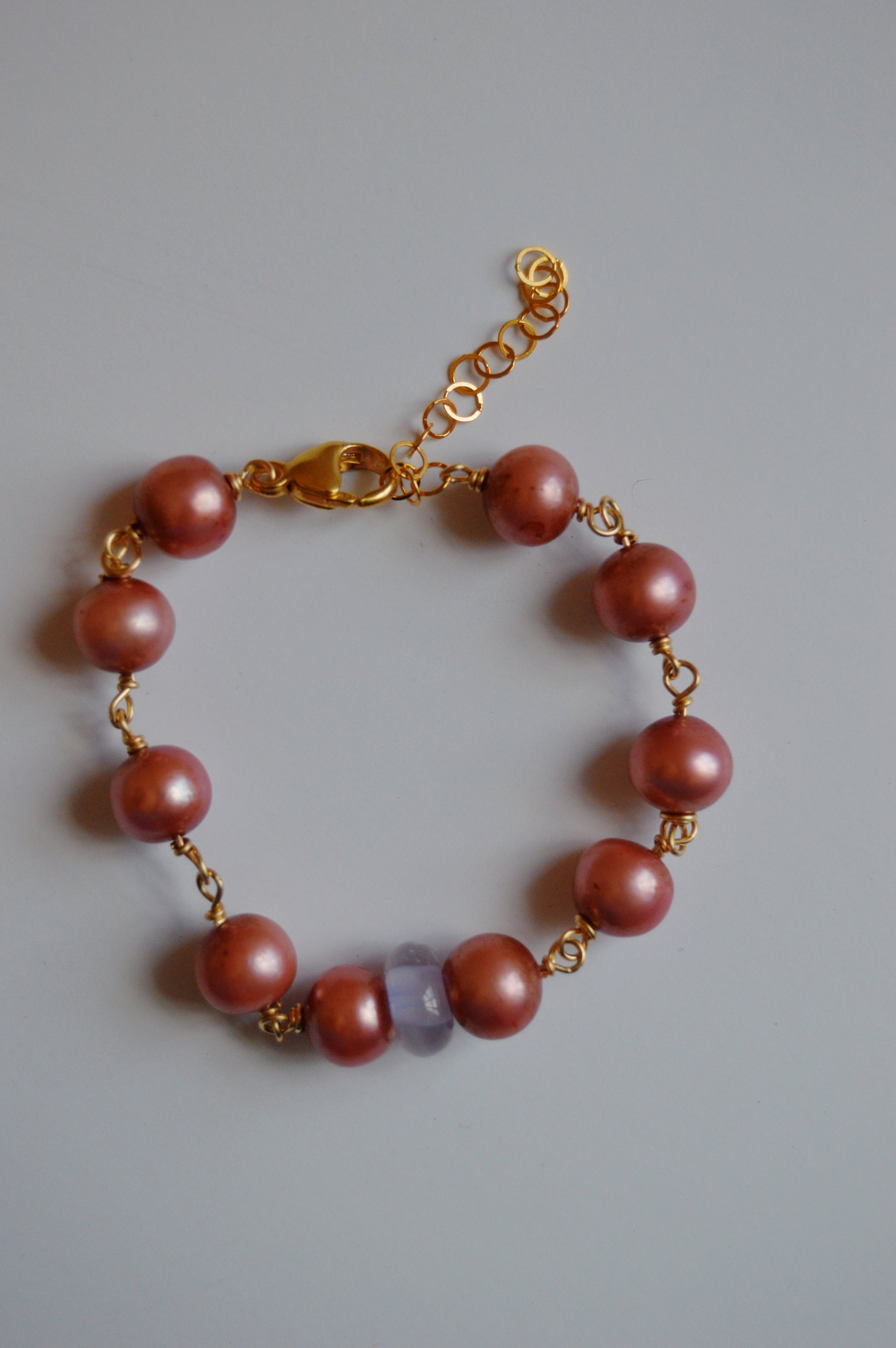 The Pearl and Glass Bracelet features pink handmade lampwork glass beads, a translucent lavender bead, and a 14k gold-filled chain with clasp, displayed on a light gray background.