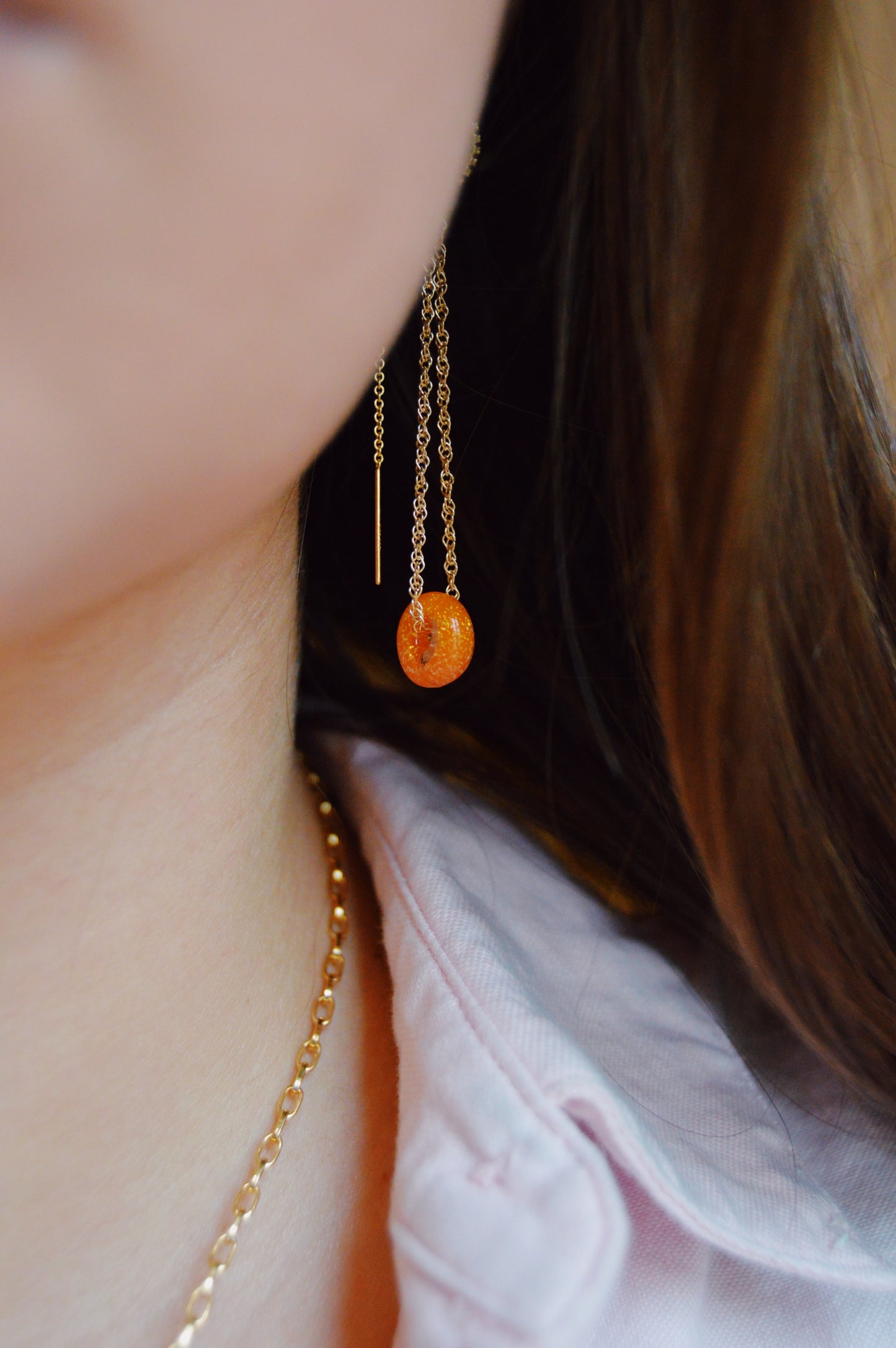 A close-up of a person wearing Donut Threaders- Sunshine Sparkle: artisan-made gold chain earrings with a small orange charm and a matching necklace, partially visible against a light pink collared shirt.
