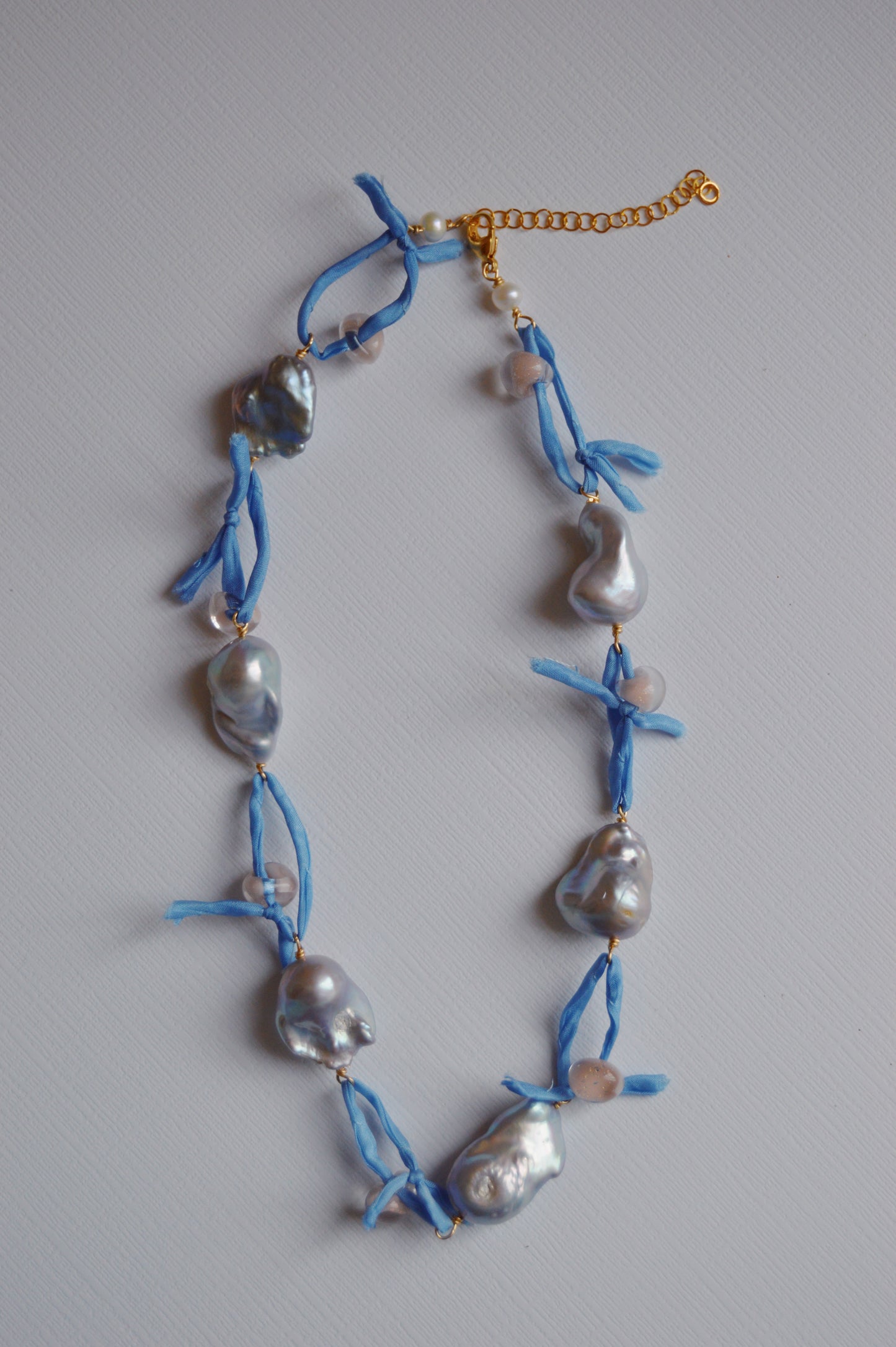 The Silk and Pearl Layer- Periwinkle showcases irregular, lustrous freshwater pearls and round beads on a blue silk ribbon with a gold chain, elegantly displayed on a white textured surface.