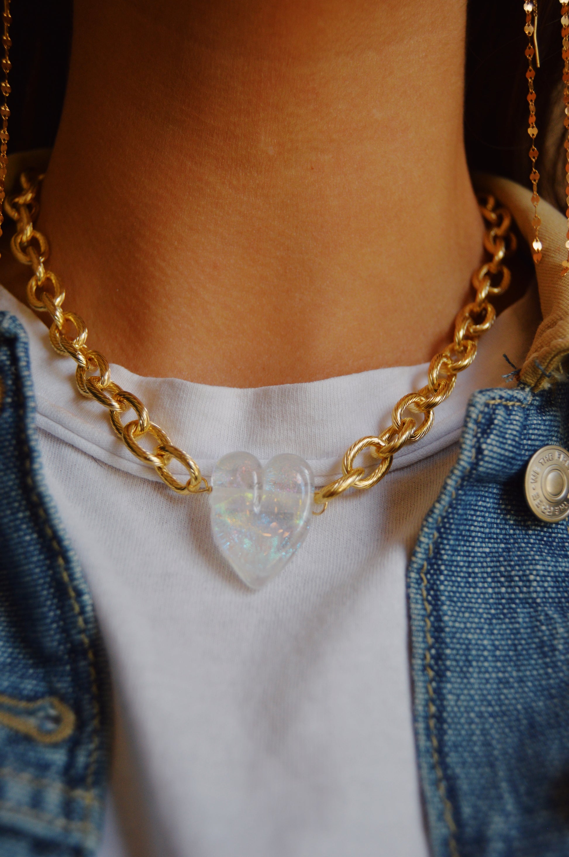 A close-up of a person wearing the Chunky Plated Glass Heart Necklace in Whites & Clears, styled with a white shirt and denim jacket.