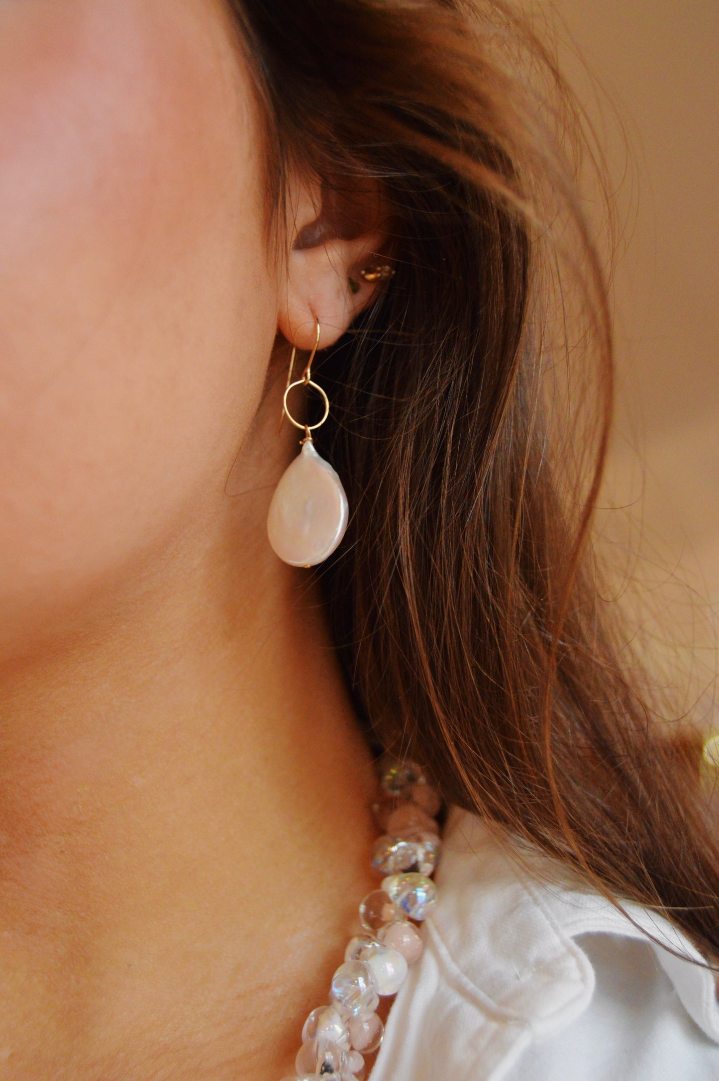 Close-up of a woman's ear wearing Pearl Drops, 14k gold-filled hoop earrings with teardrop-shaped pale gemstone pendants. She has long brown hair, a beaded necklace, and a white top, highlighting elegant handcrafted jewelry.