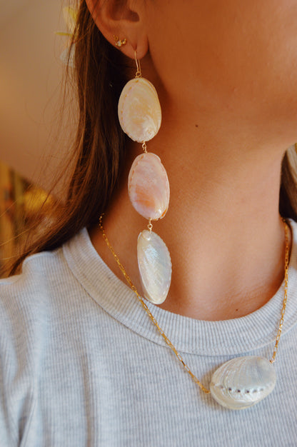 A close-up of a person wearing the Rainbow Abalone Drops—large, dangling earrings with three real abalone shells—and a matching necklace featuring a single shell on a 14k gold filled chain, paired with a light gray ribbed shirt.
