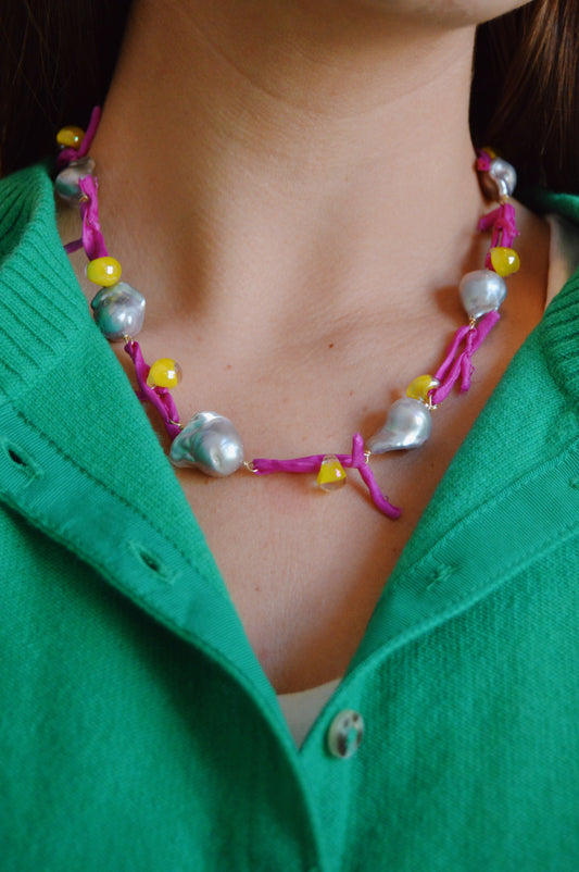 A close-up shows a person wearing a green collared sweater and the Silk and Pearl Layer- Pink Lemonade Shimmer necklace, featuring pink strands, large silver beads, and small yellow beads. The focus is on the necklace and upper chest area.