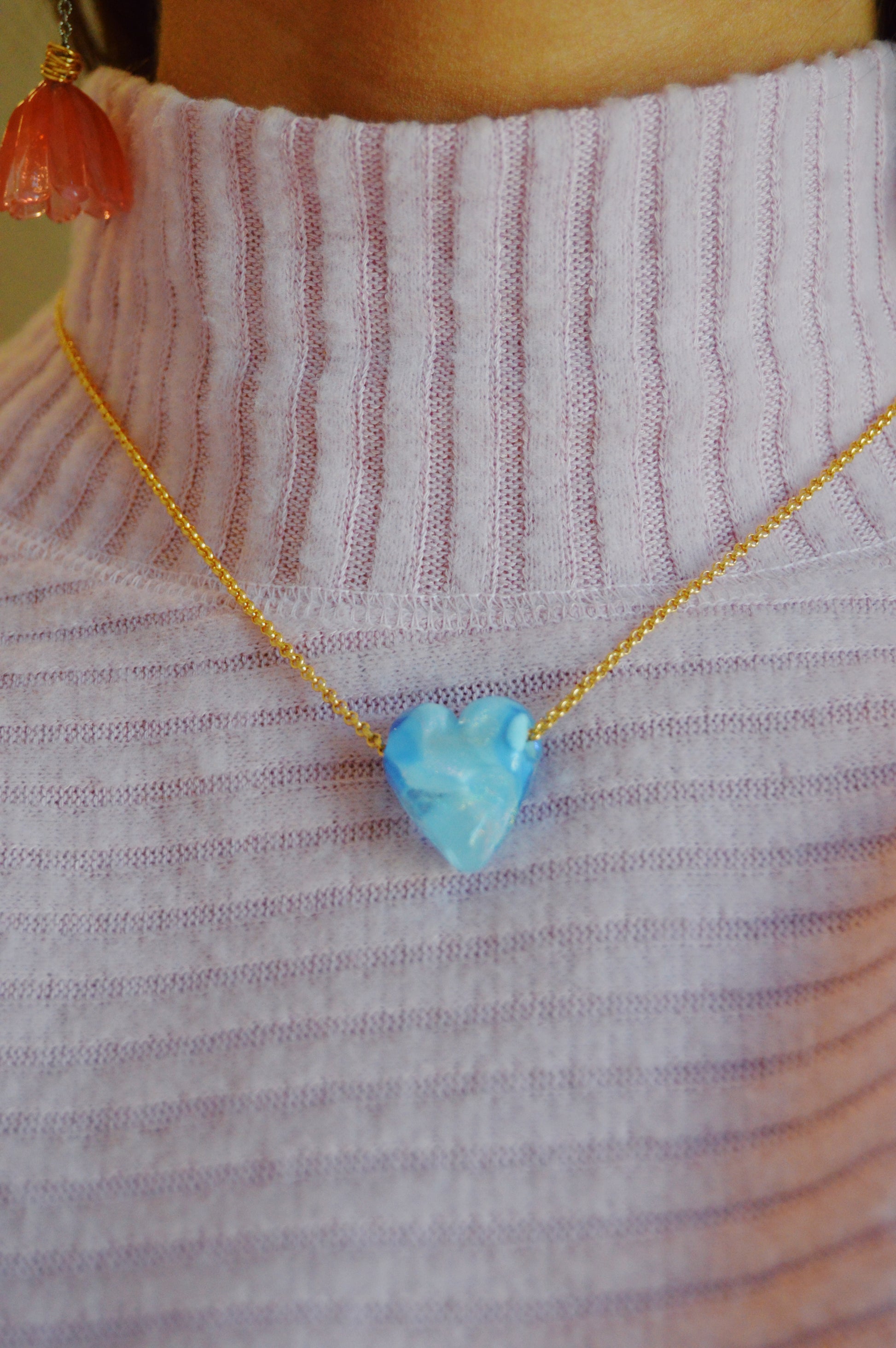 A person wearing a ribbed lavender turtleneck, a blue Glass Heart Necklace, and a pink flower-shaped earring.