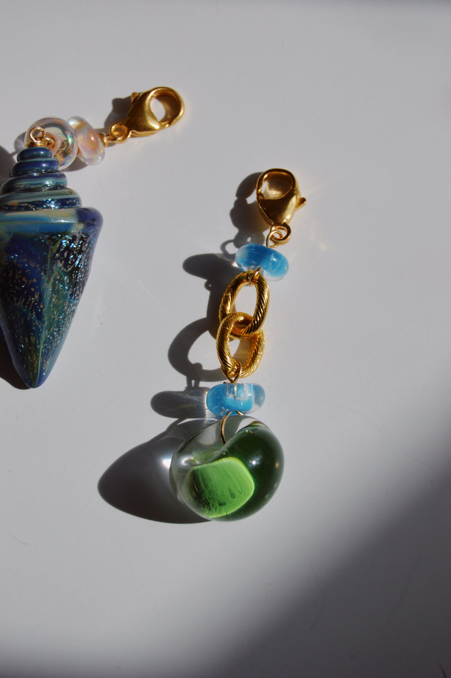 A close-up of two beaded keychains on a white surface: one features a blue cone-shaped bead with iridescent accents; the other is the Grand Bubble Charm- Green, showcasing 14k gold-filled links, blue lampwork glass beads, a green glass bead, and gold clasps.
