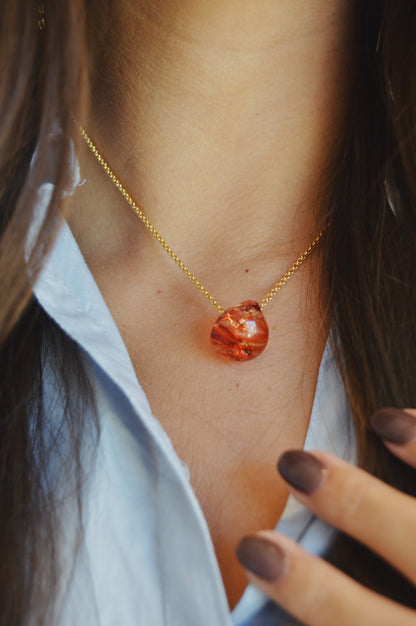 Close-up of a person wearing the Grand Bubble Single - Holiday, a gold chain necklace with an amber pendant and gold filled findings, styled with a light blue shirt. Their painted nails rest near the collar.